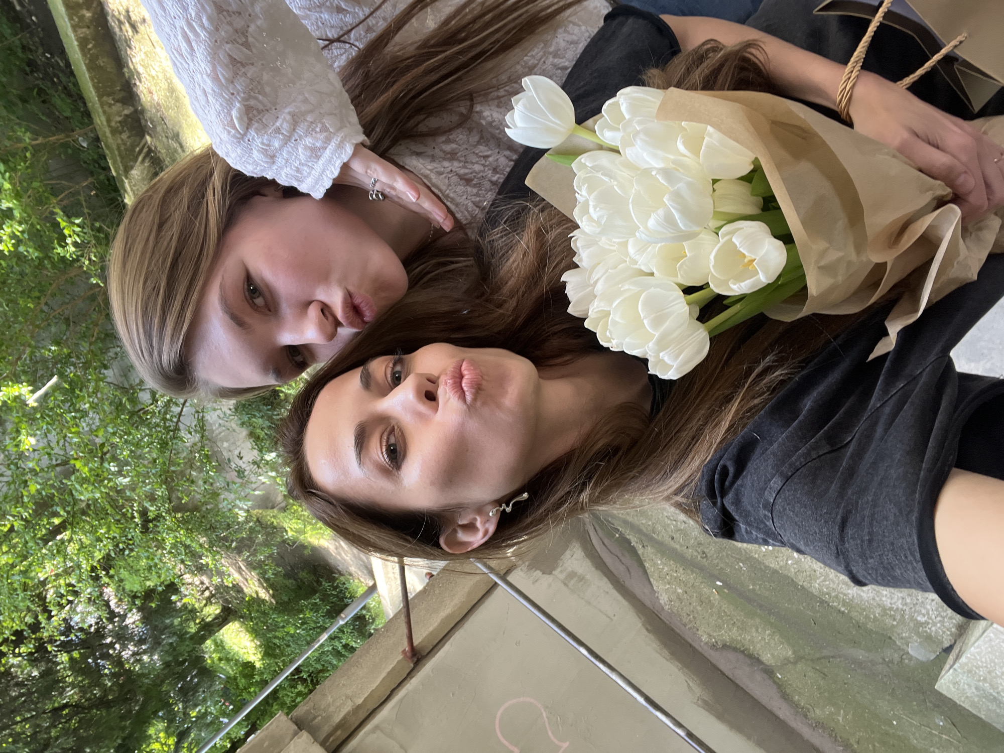 Two women pose together with puckered lips, holding a bouquet of white tulips wrapped in brown paper. The woman on top wears a white lace top. The woman below wears a dark t-shirt. They are positioned outdoors, near a concrete wall and metal railing, with trees and foliage visible in the background. The location is likely in the city of Vozdovac Urban Municipality, Serbia.