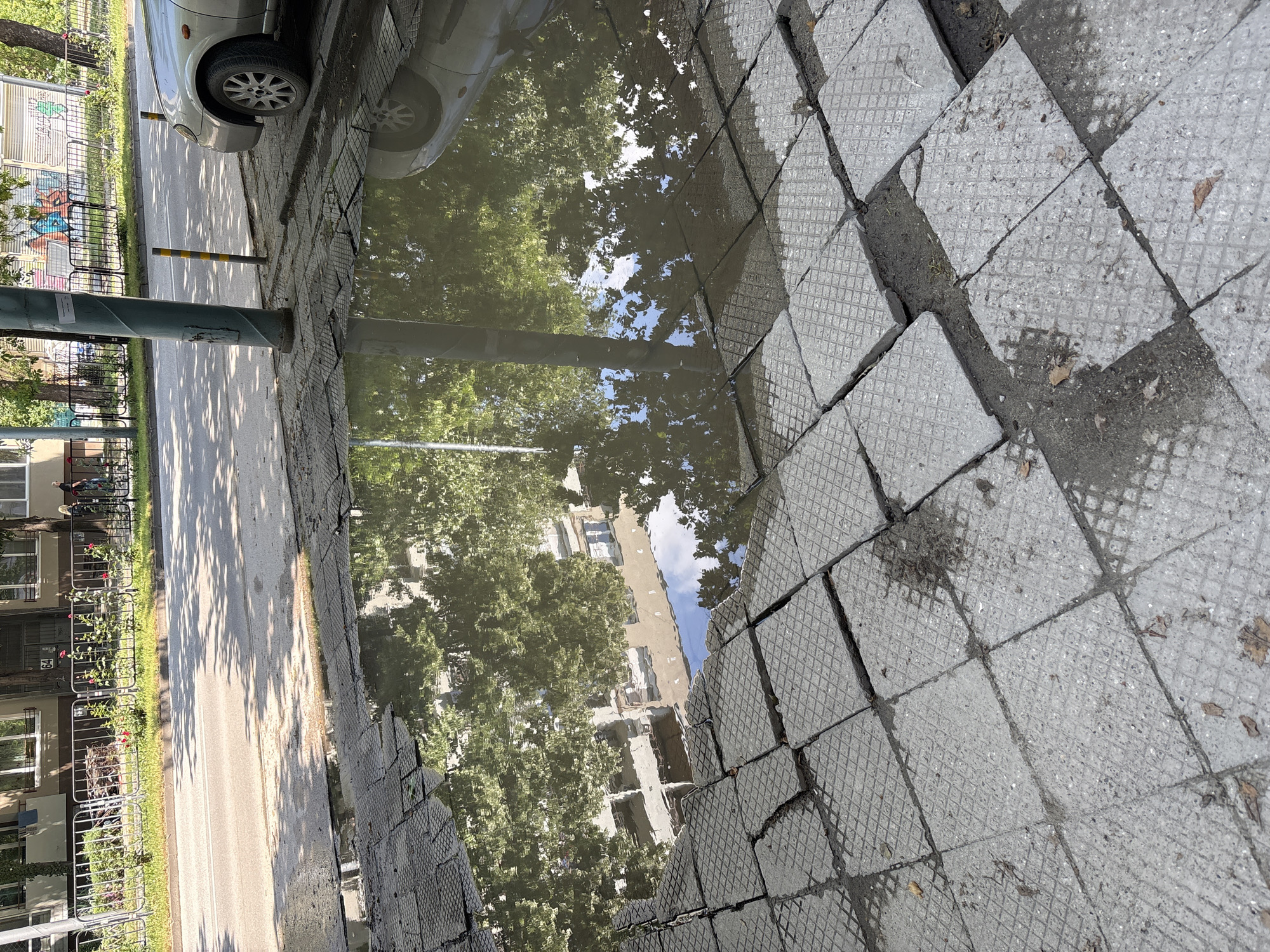 The scene is a wet street in Plovdiv, Bulgaria, reflecting the surrounding urban environment. A silver car is parked on the left edge of the image. A puddle of water on the broken pavement mirrors the trees, buildings, and sky above. A green lamppost stands between the road and the puddle. A metal fence runs along the roadside, behind which are buildings and greenery. The pavement consists of gray, patterned square tiles, many of which are cracked or displaced.