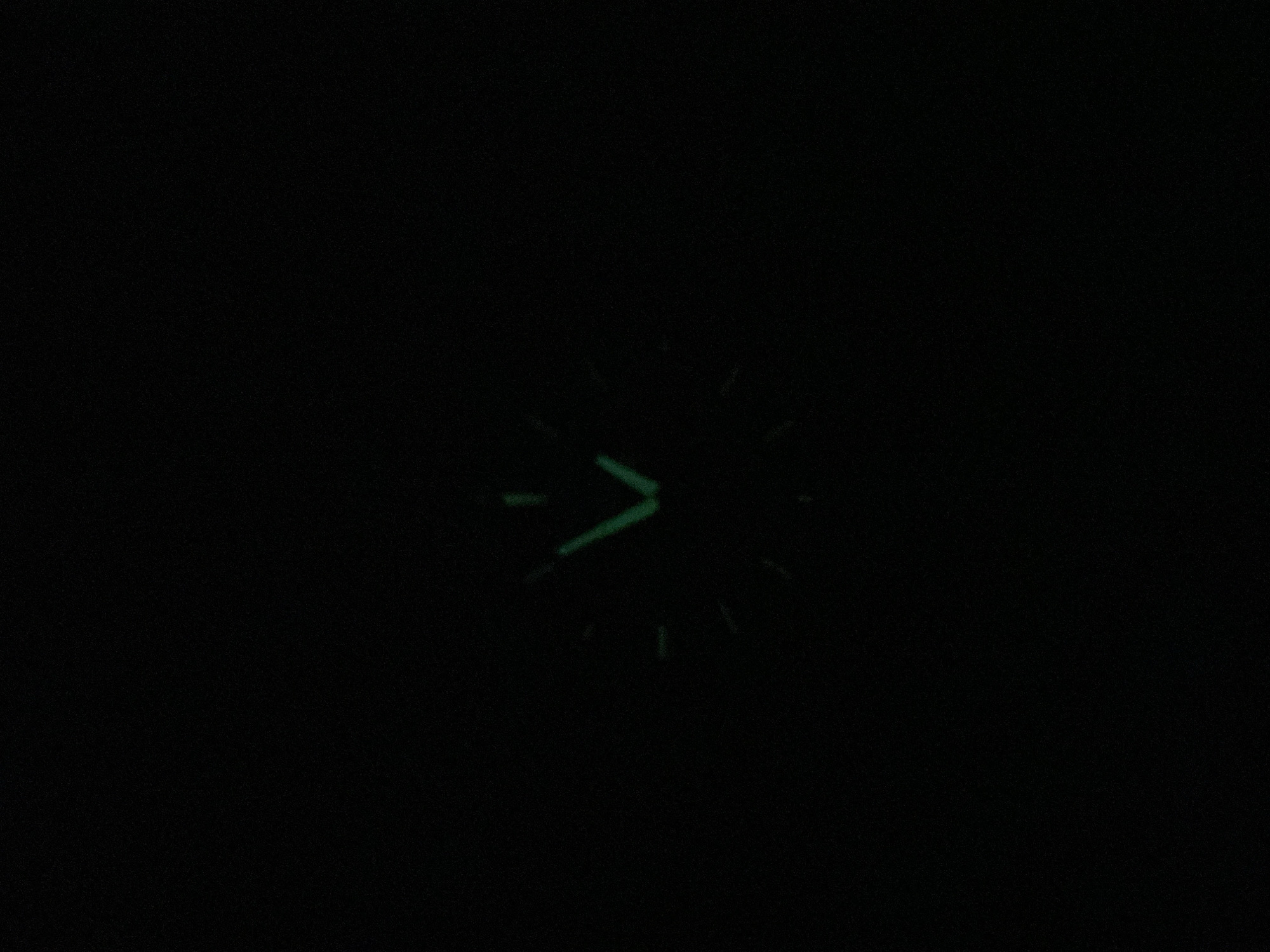 A close-up image of a watch face with luminous hands and hour markers is captured in near-total darkness. The hour hand is slightly past the 6 position, while the minute hand is near the 11 position. The image was taken in an unknown location in the city of Unknown, Unknown.