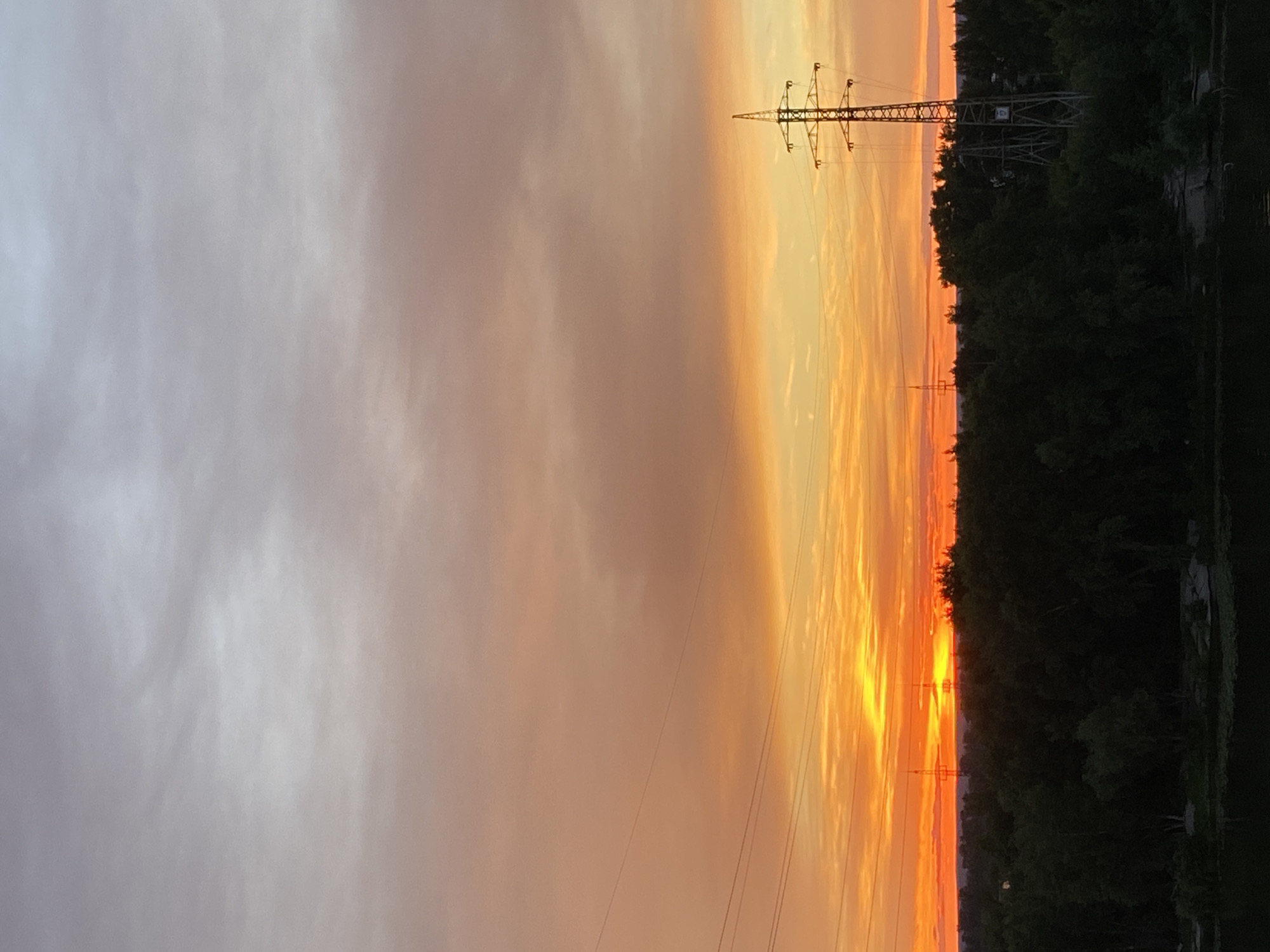 The media file depicts a sunset over a landscape in Novokuybyshevsk, Russia. The scene is dominated by a dramatic sky transitioning from pale grey clouds on the left to vibrant hues of orange and yellow near the horizon on the right, indicating the time of day is sunset. A large electricity pylon stands prominently against the sky, with power lines stretching across the frame. To the right of the pylon, a dense line of dark green trees forms a stark contrast against the colorful sky, suggesting a wooded area bordering the scene. In the far right, a body of dark water is visible, possibly a river or lake, with some indistinct structures along its edge. The overall mood is tranquil and serene, characteristic of a natural sunset. No people are visible in the image, and there is no visible text.