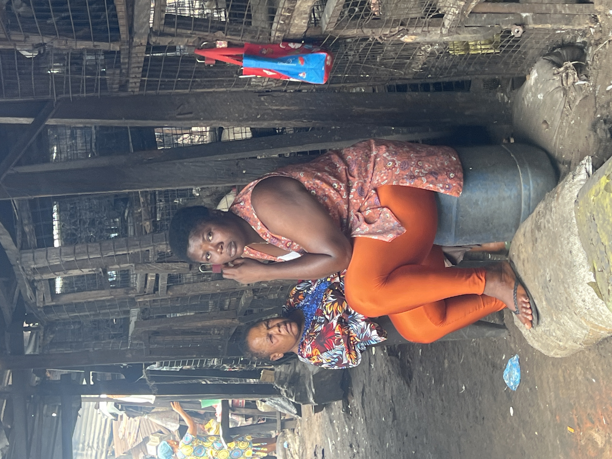 In a marketplace in Nkpor, Nigeria, two women are visible. The woman in the foreground is seated on a blue plastic barrel, wearing an orange sleeveless dress with a floral print and bright orange leggings. She has her right hand raised to her ear. The second woman is seated to her left on a rough, circular stone, wearing a colorful dress with a blue neckline and a floral pattern of red, yellow, and blue. She is looking towards the left. The environment appears to be outdoors, under a dilapidated structure with wooden beams and a wire mesh enclosure. A red bag with a blue design is hanging on the mesh. The ground is dirt and debris, with some plastic waste visible. In the background, more market stalls and structures are present.