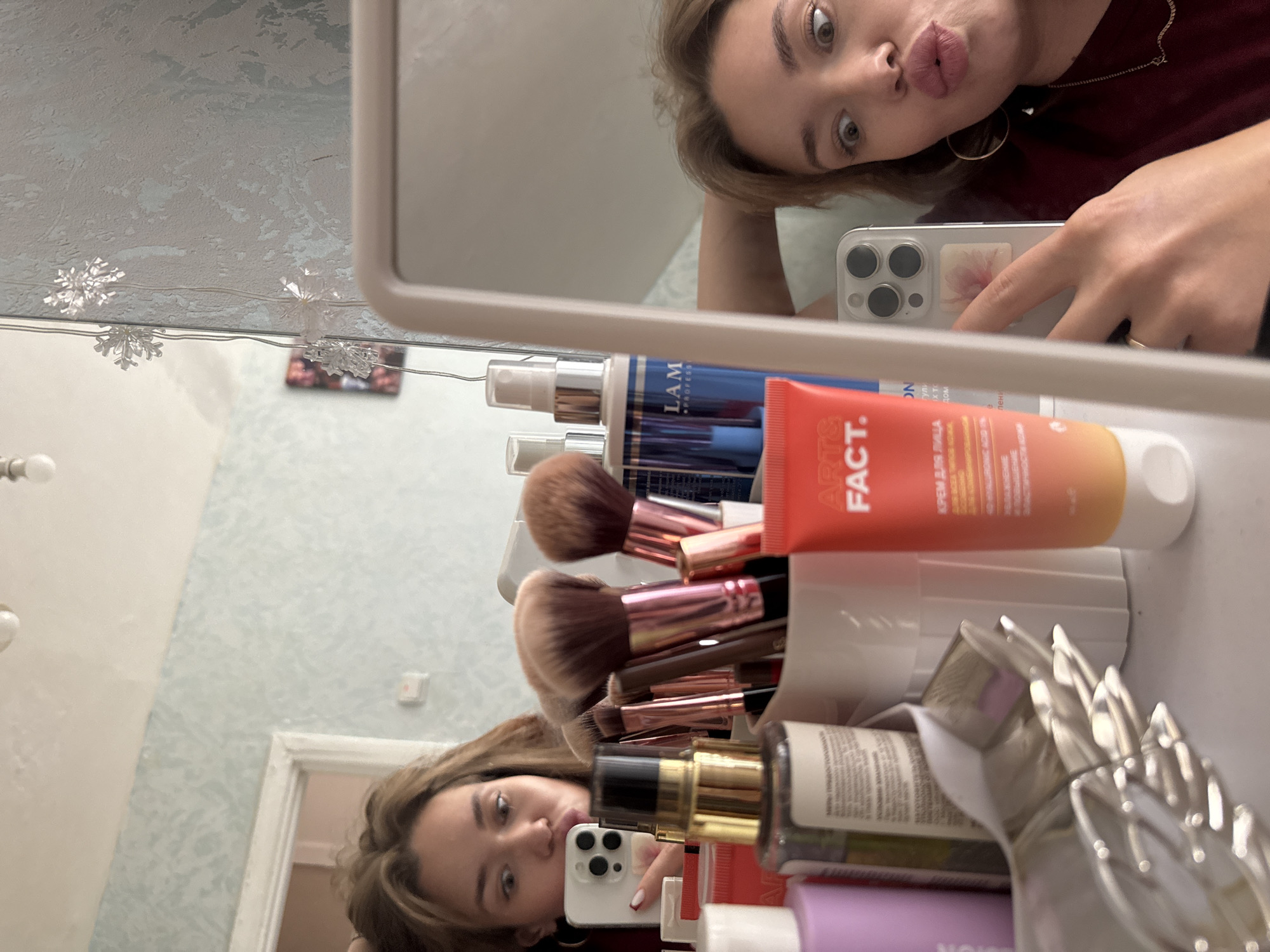 A young woman is taking a selfie in a mirror, with a collection of makeup and beauty products in front of her. The woman has light brown hair and is looking at the camera with a playful expression, puckering her lips. She is wearing a dark red top and large hoop earrings. Her right hand is holding a silver smartphone, which is capturing the reflection.
In the foreground, a white container holds various makeup brushes with pink and gold handles. Behind the brushes, a red tube labeled "ART FACT" is visible, along with a blue bottle with a silver pump that says "LAM." Other beauty products are also present, including bottles and tubes.
The background is a wall with a subtle pattern, and there are string lights with snowflake shapes hanging across the top of the mirror. The overall scene appears to be in a domestic setting, possibly a vanity or dressing area. The lighting suggests it is daytime or a well-lit indoor space. The reflection in the mirror also shows a doorway in the background.