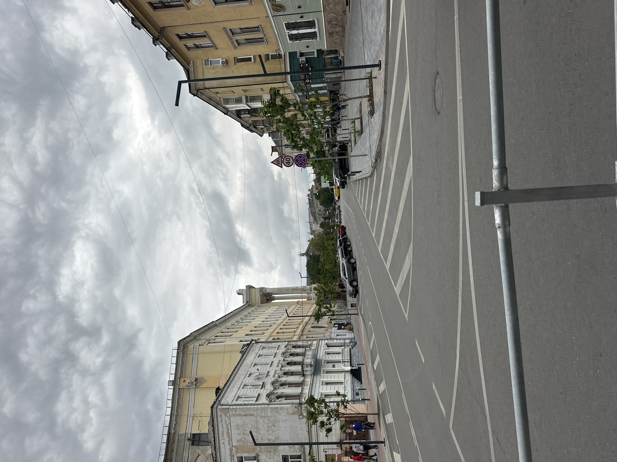 This image captures a street scene in Ternopil, Ukraine, featuring an intersection with a pedestrian crossing. On the left, a weathered, light-colored building stands adjacent to a larger, yellow building with classical architectural elements. The street itself is paved and marked with white lines for traffic lanes and a pedestrian crossing. Several cars are parked along the right side of the street, which is lined with trees and multi-story buildings in various architectural styles. Traffic signs, including a speed limit sign (40) and a prohibition sign, are visible. The sky is overcast with dark, textured clouds, suggesting a cloudy day. A metal barrier is visible in the foreground. Several pedestrians are walking on the sidewalks.