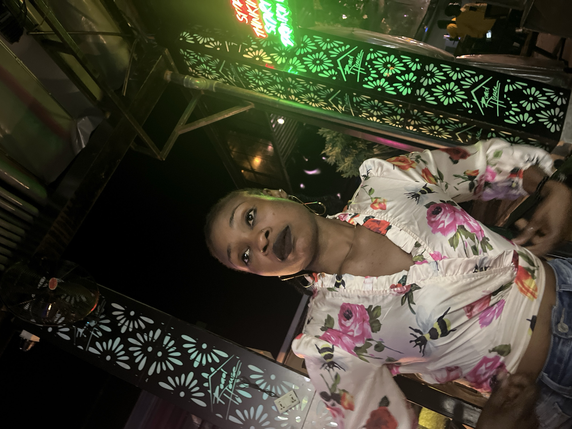A low-angle, full-body shot captures a young Black woman from the chest up, posing outdoors at night. She has dark skin, dark hair pulled back into a bun, and wears dark lipstick. She is dressed in a cream-colored, floral-print blouse with pink and red flowers and bees, featuring a ruffled V-neck and puffed sleeves. Her ears are adorned with gold hoop earrings. Her body is angled slightly to her right, and her gaze is directed upwards and to her left.
Behind her, on her left, a black pillar with a laser-cut daisy pattern glows with a pale turquoise light. A ceiling fan is visible above and to the left. To her right, another black pillar with the same daisy pattern is illuminated by bright green neon lights. The neon sign reads, "STILL THINKING START DRINKING," and the "Road House" logo is visible on both pillars. The environment appears to be an outdoor establishment, possibly a bar or restaurant, with structural elements and lighting suggesting an urban setting at night. The background is dark, with hints of greenery and other illuminated structures, suggesting a lively atmosphere.