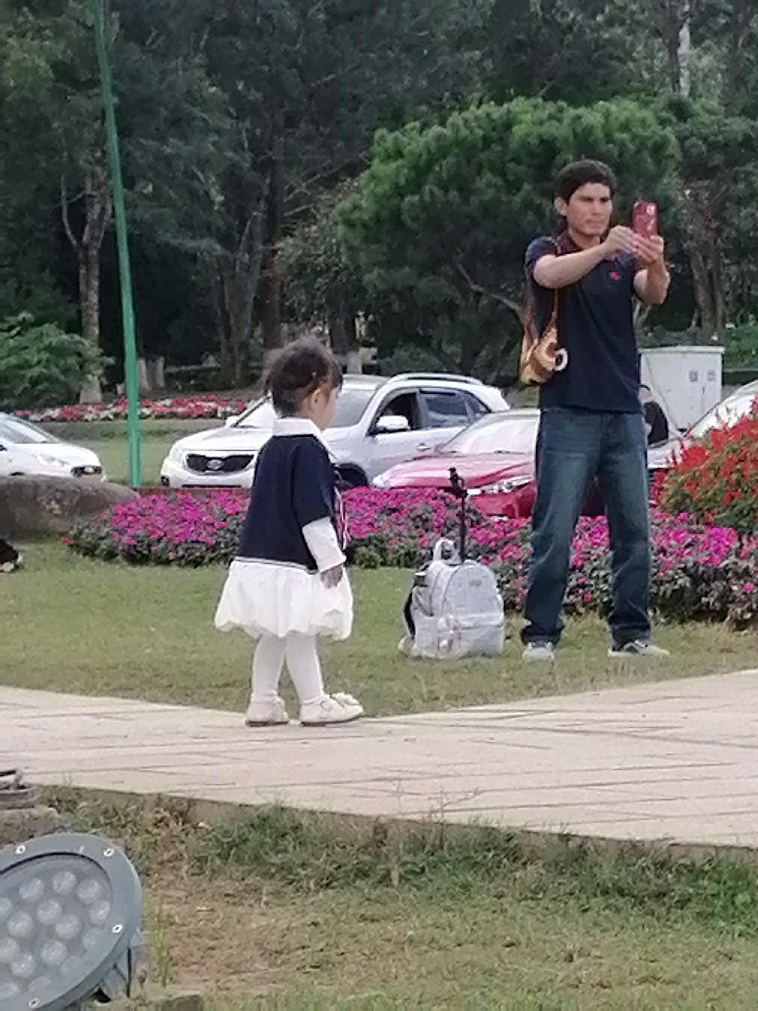 In Da Lat, Vietnam, a young girl wearing a dark blue top, white dress, and white leggings stands in a park, facing away from the camera. She appears to be walking on a paved path. To her right, a man in a dark blue polo shirt and blue jeans is taking a photo with his red smartphone. He has a patterned bag slung over his shoulder. A light gray backpack with a black strap is on the ground near him. In the background, several cars are parked, and a patch of bright pink flowers is visible. The setting is a park with lush green grass and trees. The lighting suggests it is daytime, possibly late afternoon or early evening, with overcast weather. There is no visible text in the image.