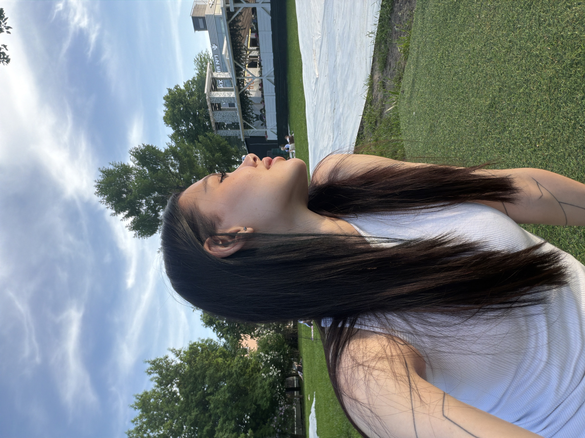 A woman with long, dark hair lies on her back in a grassy area. She is wearing a white tank top and has a tattoo on her arm. Her head is tilted back, and she is looking upward. In the background, there are trees, a white sheet, and a building with a metal framework. The sky is blue with white clouds. The scene is located in Moscow, Russia.