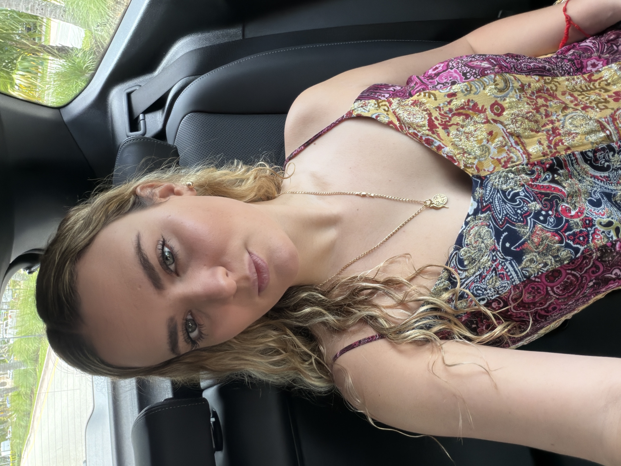 A young woman is reclined in a black car seat, presumably in the back of the car, as indicated by the positioning and surrounding interior features. She is wearing a patterned top with thin straps, a gold necklace, and a red bracelet on her arm. Her hair is blonde and curly, and she is looking toward the camera with a neutral expression. Outside the car window, green foliage is visible, suggesting an outdoor, potentially tropical or subtropical environment. Based on the prompt, the location is Key Biscayne, United States of America.