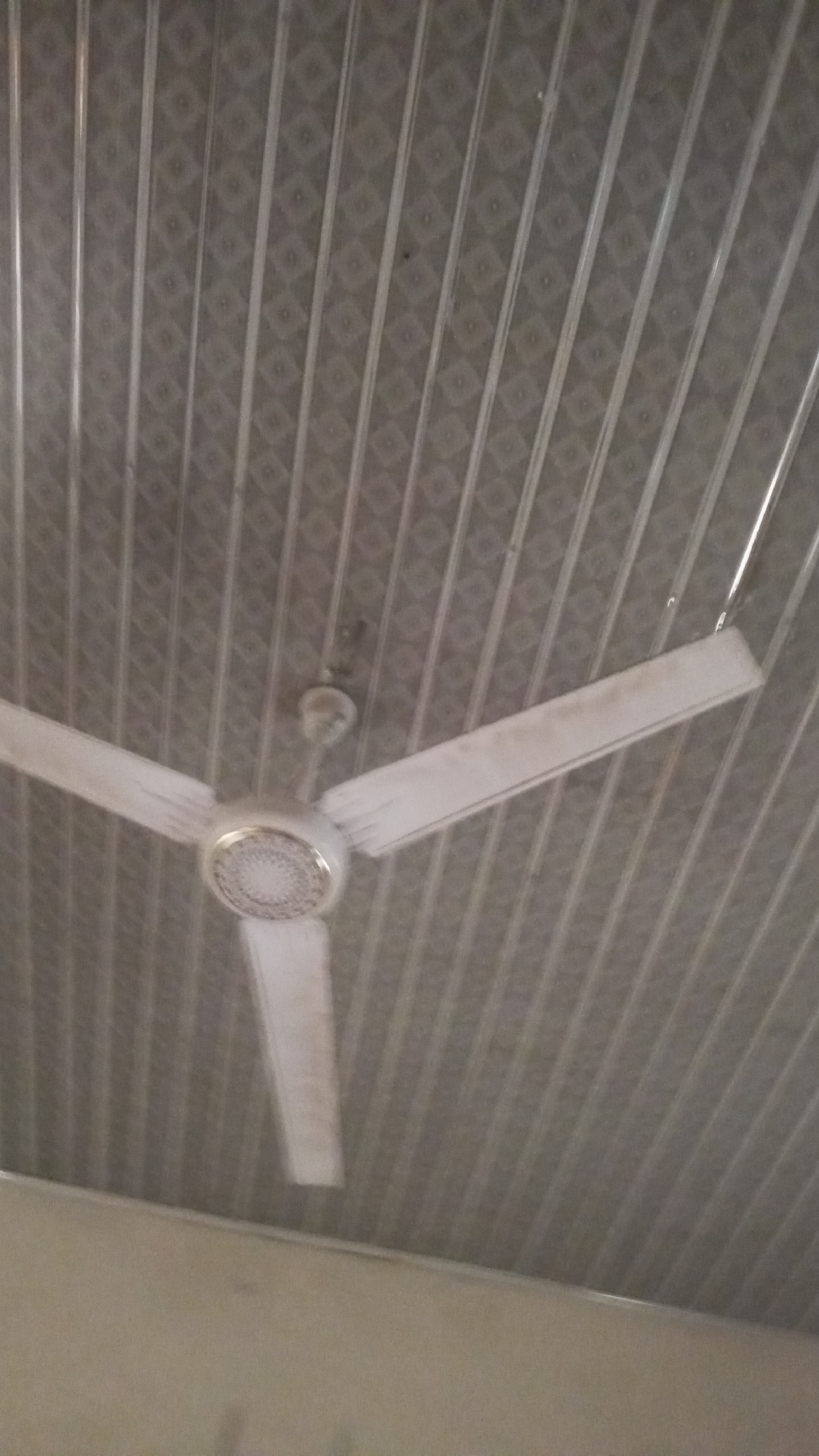 A white ceiling fan with three blades is in motion, suspended from a gray, patterned ceiling. The fan's central housing has a circular, textured design. The ceiling is composed of long, parallel panels with a repeating diamond-shaped pattern. The bottom of the image shows a section of a lighter-colored wall or structure. The scene is indoors, and based on the provided context, it is located in Jalingo, Nigeria.