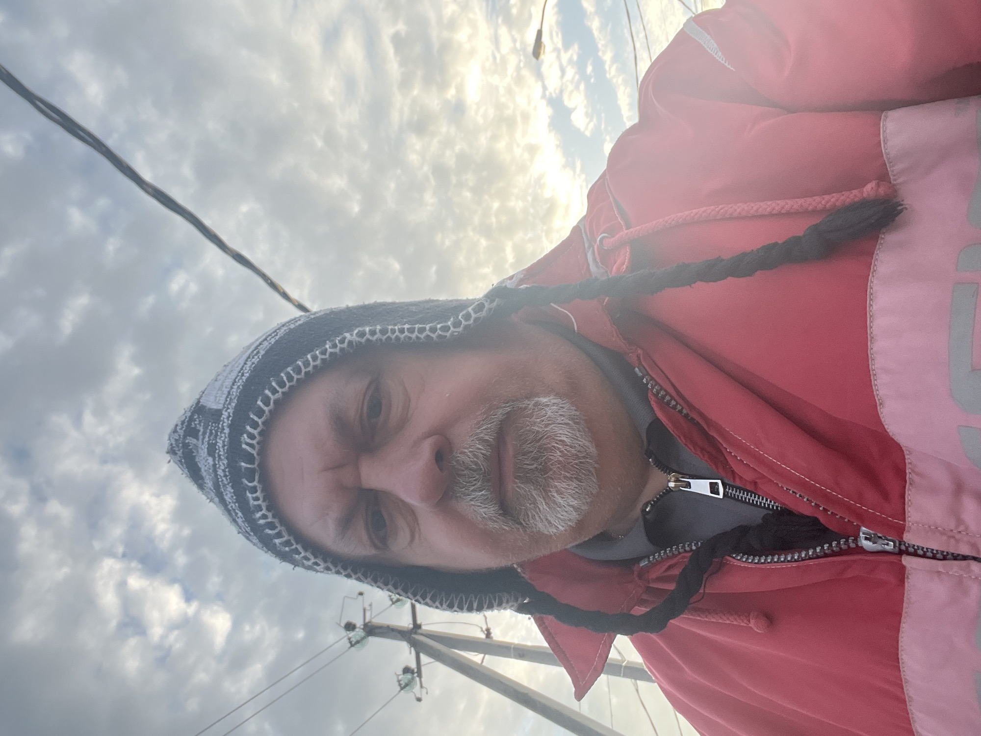 A man is seen from a low angle, wearing a patterned knit hat and a red and light pink jacket. The zipper of the jacket is visible. He has a gray beard and mustache. The background includes a cloudy sky, utility poles, and overhead wires. The location cannot be determined as Podolsk, Russia from this image alone.