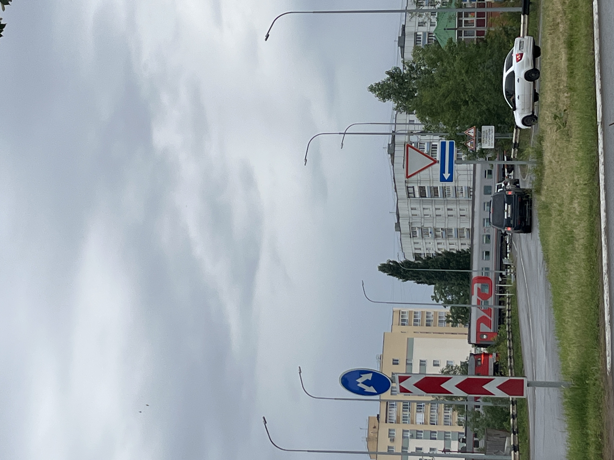 The image depicts an urban or suburban street scene under an overcast sky, suggesting a cloudy day. The road appears wet, possibly from recent rain, running parallel to a set of train tracks on the left side of the image. Residential buildings of varying heights and colors, interspersed with green trees, line the background.
On the right side of the road, a white hatchback car is parked on a grass verge. Further down the road, a dark sedan or SUV is visible on the asphalt. A passenger train, displaying the distinctive red "РЖД" (RZhD - Russian Railways) logo, is on the tracks.
Several traffic signs are prominent. A red-bordered "Give Way" (inverted triangle) sign is visible near the parked white car. Below it, a blue rectangular sign with a white double-headed arrow pointing downwards indicates two-way traffic. Another white rectangular sign nearby has Russian text, partially readable as "ОТДОХНИ ПЕРЕД БУГРОВОЙ АВТОМОЙКОЙ," which translates to "Rest before Bugrovaya Car Wash," suggesting a nearby business. Further along the road, a blue circular sign with two white wavy arrows pointing left and right likely indicates "Pass on both sides" or an obstacle bypass. Adjacent to this, a red and white chevron sign with arrows pointing right signifies a sharp turn or obstacle.
The scene captures a typical, mundane daytime street view in an area with both road and rail transport.