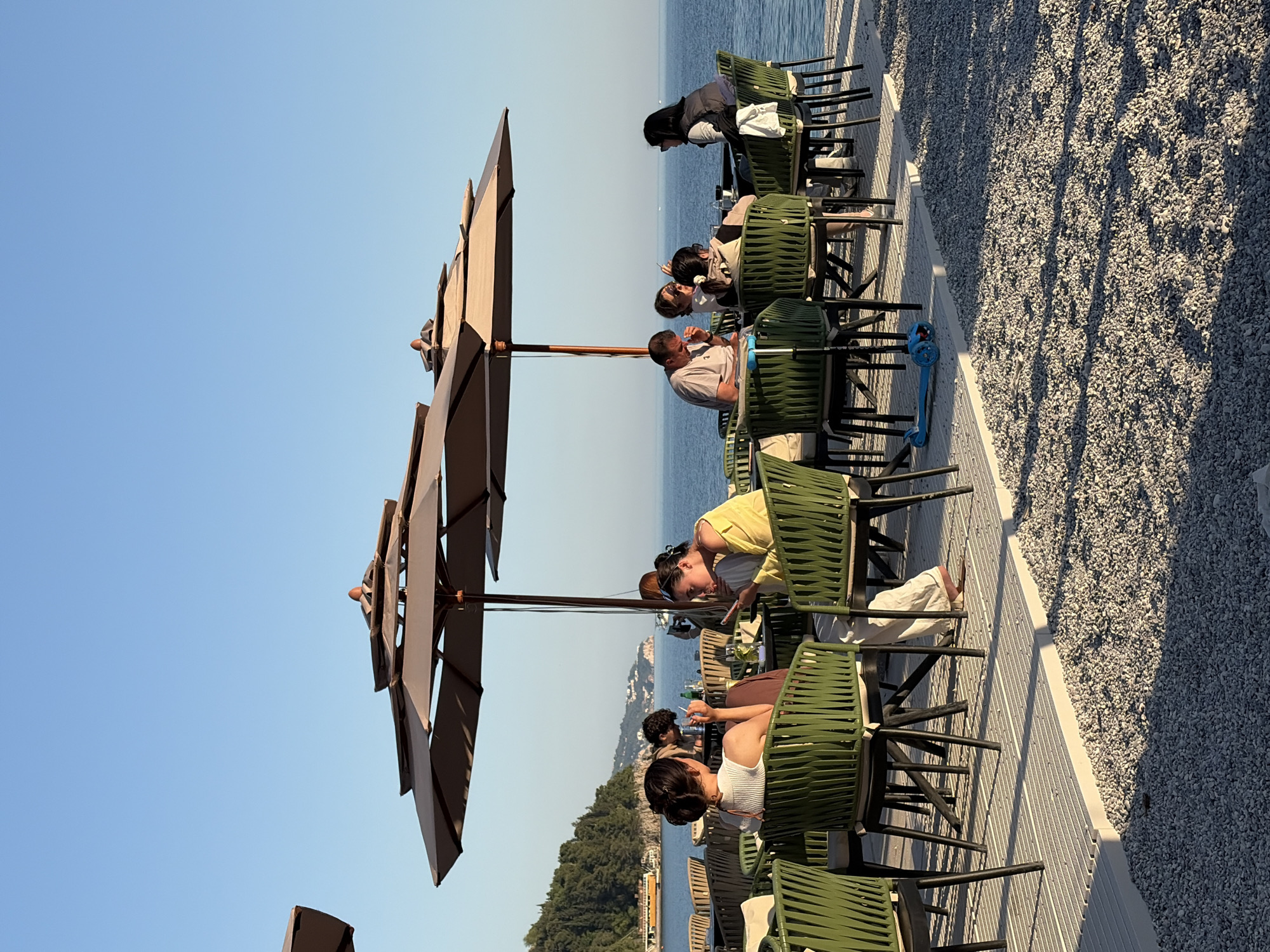 The image captures an outdoor cafe or restaurant situated along a waterfront, likely in the late afternoon or early evening due to the long shadows cast across the ground. The setting features a clear, bright blue sky and calm, blue sea stretching to the horizon, with a green, tree-covered coastline visible in the distance.
In the foreground and midground, multiple groups of people are seated at tables under large, light-brown patio umbrellas. The seating consists of green, wicker-style chairs and dark-colored tables. People are casually dressed, engaged in conversation, and appear to be relaxing and socializing. One person in a white top and another in a yellow top are visible leaning forward, looking at something on a table. Further along, a man in a light polo shirt is seated, and another person in a black jacket and head covering is looking towards the water.
The cafe area is on a paved or boardwalk-like surface that runs parallel to the water. Adjacent to this seating area is a strip of light-colored gravel or small stones, across which pronounced shadows of the chairs and umbrellas are cast, indicating the sun is low in the sky. A blue child's scooter is parked next to one of the tables. No visible text is present in the image.