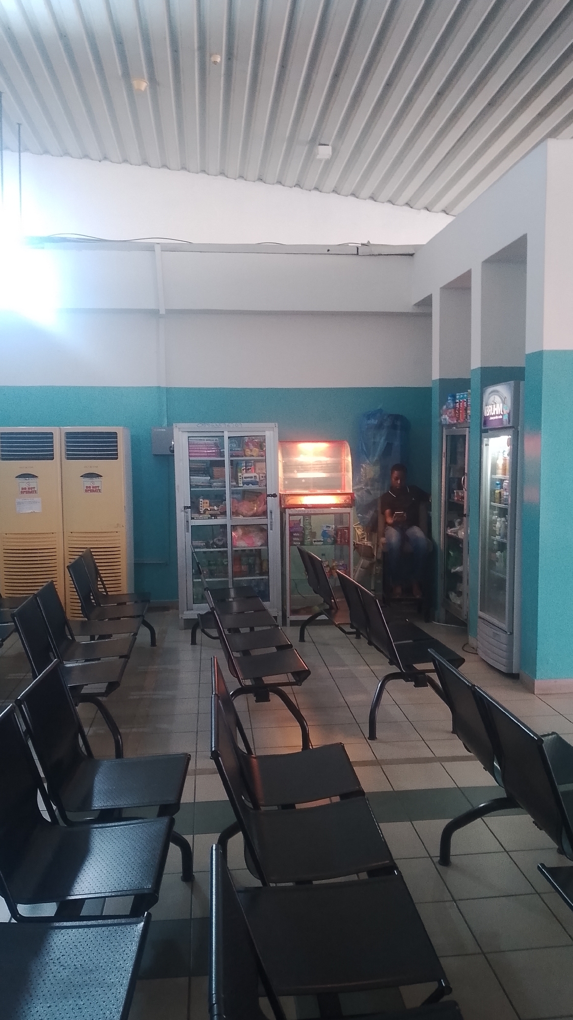 The image shows an interior space, likely a waiting area. Rows of dark metal chairs are arranged on a tiled floor. Along the back wall are two tall, light-colored air conditioning units, a display case stocked with various items, a heated display case, and a refrigerated drink case. A person is seated near the refrigerated drink case, looking at a mobile device. The walls are painted in two tones, white at the top and turquoise below, separated by a dark stripe. The ceiling is corrugated metal.
Location is unknown.