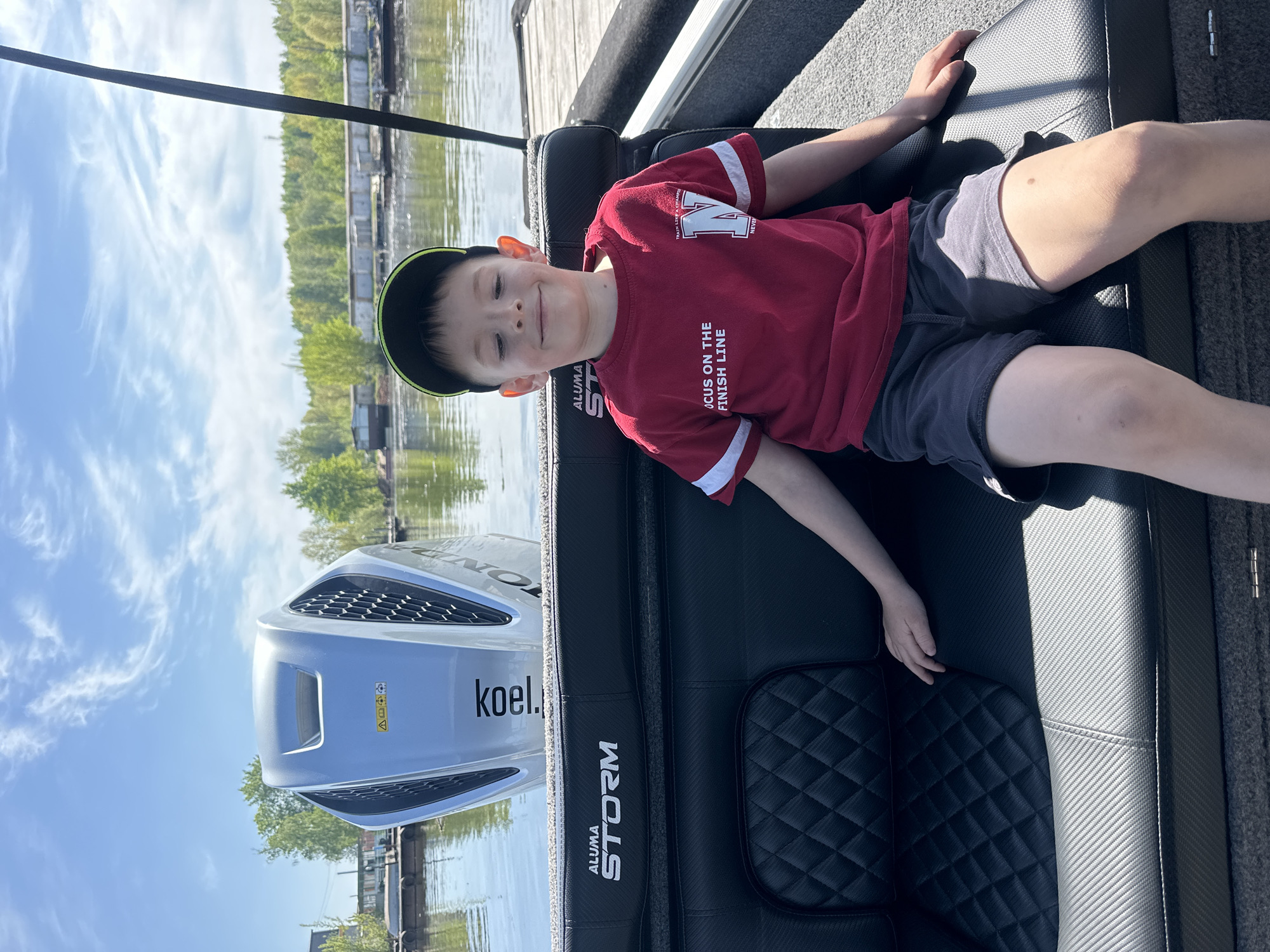 A boy is lying on the seat of a boat. He is wearing a red shirt with white sleeves, gray shorts, and a baseball cap. The boat is on a body of water surrounded by trees and buildings. The name "ALUMA STORM" is visible on the side of the boat. The location is the city of Chaikovsky, Russia.