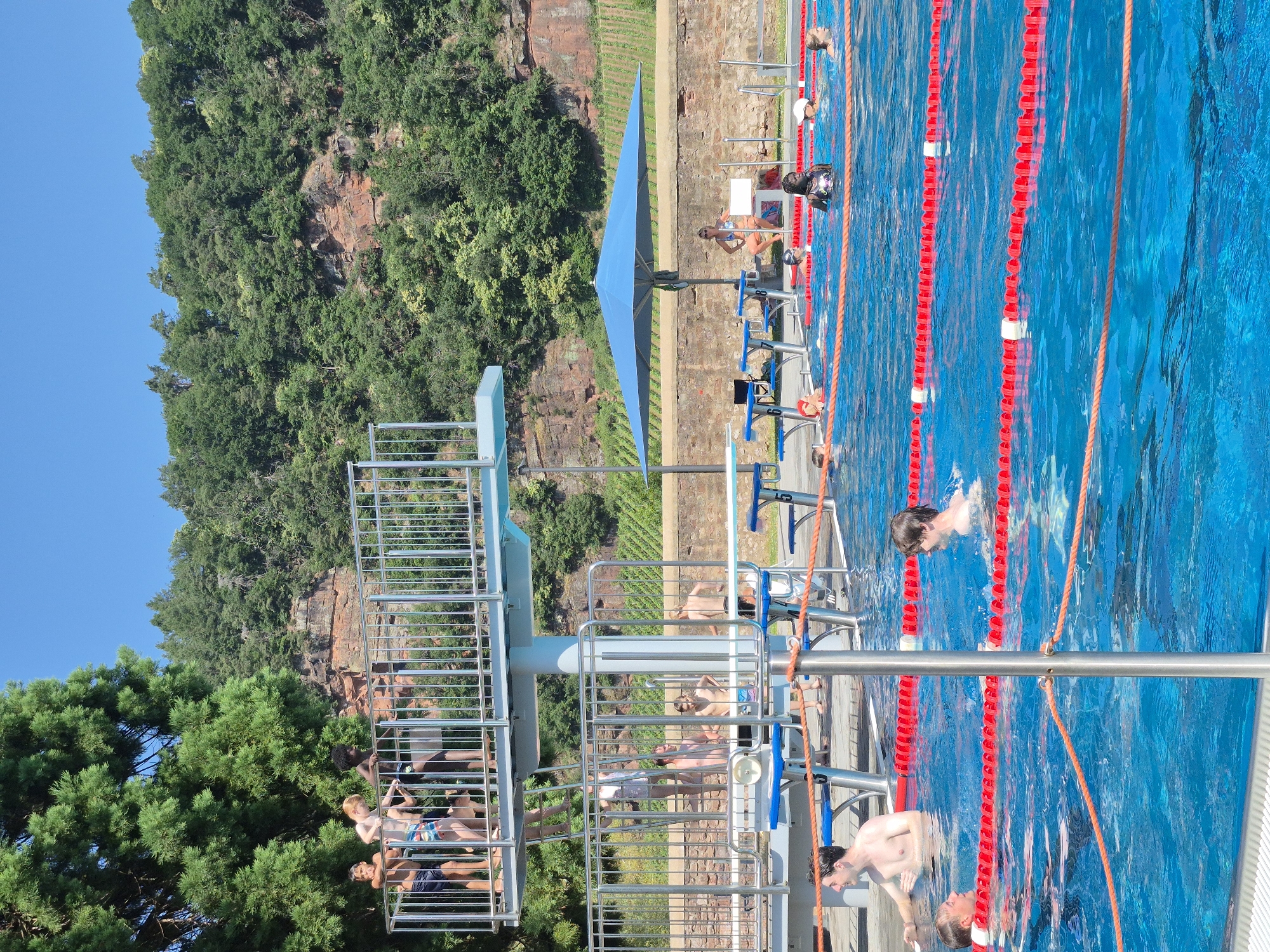 This is a medium shot, eye-level, outdoor photograph of a crowded swimming pool area on a sunny day. In the foreground, the bright blue water of the pool is divided by orange ropes and red and white lane markers. Several people are swimming in the pool, including two young men in the mid-ground and a group of children further back. In the mid-ground, there is a diving platform with several individuals standing on it and climbing a ladder. Behind the pool, a stone wall runs the length of the scene, and above that, a steep, tree-covered hillside with visible rock formations rises into a clear blue sky. A large blue umbrella provides shade for a woman sitting on a bench near the stone wall. The overall atmosphere suggests a recreational setting.