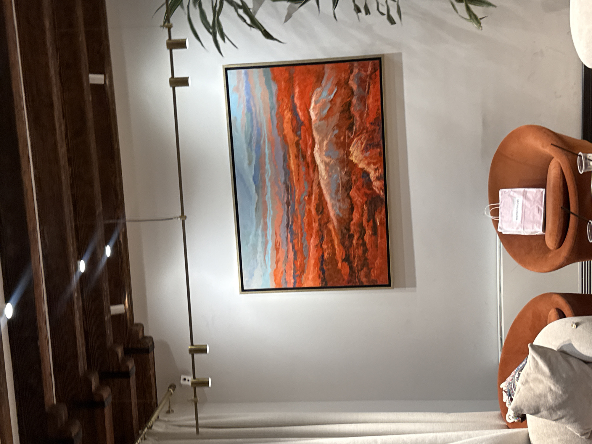 The image depicts an interior scene featuring a large, framed abstract painting of a landscape with vibrant red and orange hues, hanging on a white wall. Above the painting, a track lighting system with multiple adjustable spotlights is mounted on a dark wooden beam ceiling. To the left of the painting, a light-colored curtain hangs, and to the right, the foliage of a large plant is visible. In the foreground, two rust-colored, curved armchairs are present. One armchair has a white shopping bag with the "Women'secret" logo on it placed on the seat. Another armchair on the left has decorative pillows. This scene is located in Sasunik, Armenia.