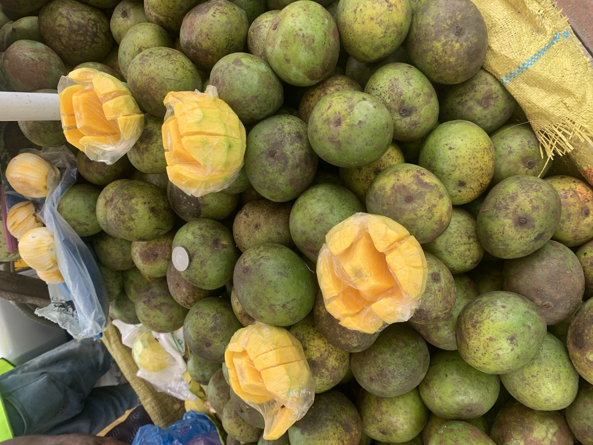 The image depicts a close-up view of a street market stall in Dar es-Salaam, Tanzania, laden with a large quantity of unpeeled, green-skinned mangoes. The fruits display natural blemishes and mottling. Interspersed within the pile are four bright yellow, ripe mangoes that have been cut into a "hedgehog" style (cross-hatched to expose the flesh) and are each enclosed in clear plastic bags, suggesting they are prepared samples or ready-to-eat portions. A silver-colored coin rests on one of the green mangoes in the upper middle section of the frame. The mangoes are contained within a yellow woven sack, partially visible at the bottom right. In the upper left background, parts of a person wearing blue jeans and a neon yellow vest are visible, standing near a light-colored vehicle. A blue plastic bag and additional lighter-skinned fruits in clear plastic are also discernible in the upper left. A vertical white pole, likely a support structure for a stall, is present in the upper center. The scene conveys a bustling outdoor commercial environment focused on fruit sales.