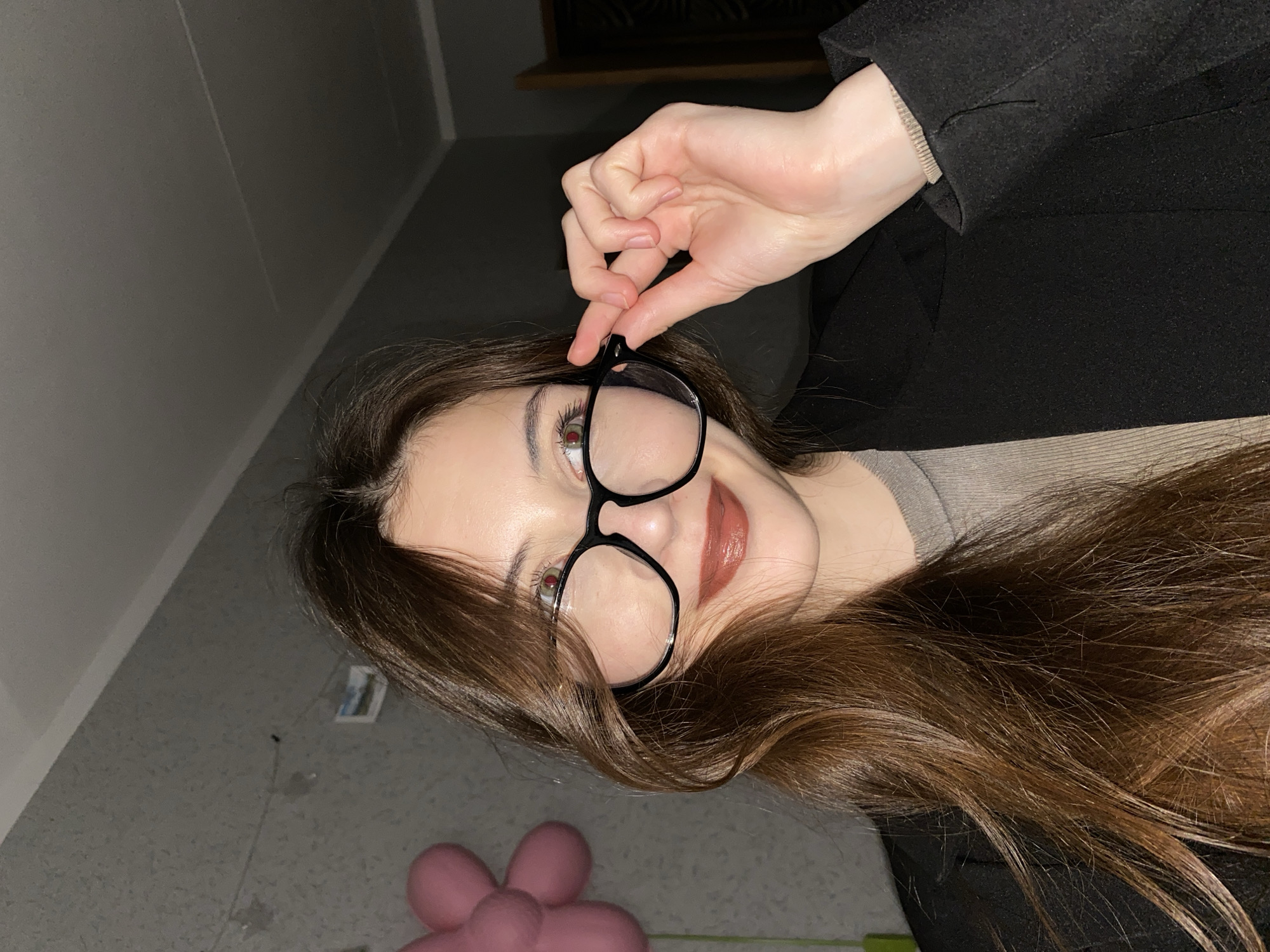 A woman with long, brown hair is holding up a pair of black-framed eyeglasses, partially obscuring her face. She is wearing a gray shirt and a black jacket. The background includes a wall, a section of flooring, and a pink flower-shaped object on the floor. There is no indication of the location being in Votkinsk, Russia.