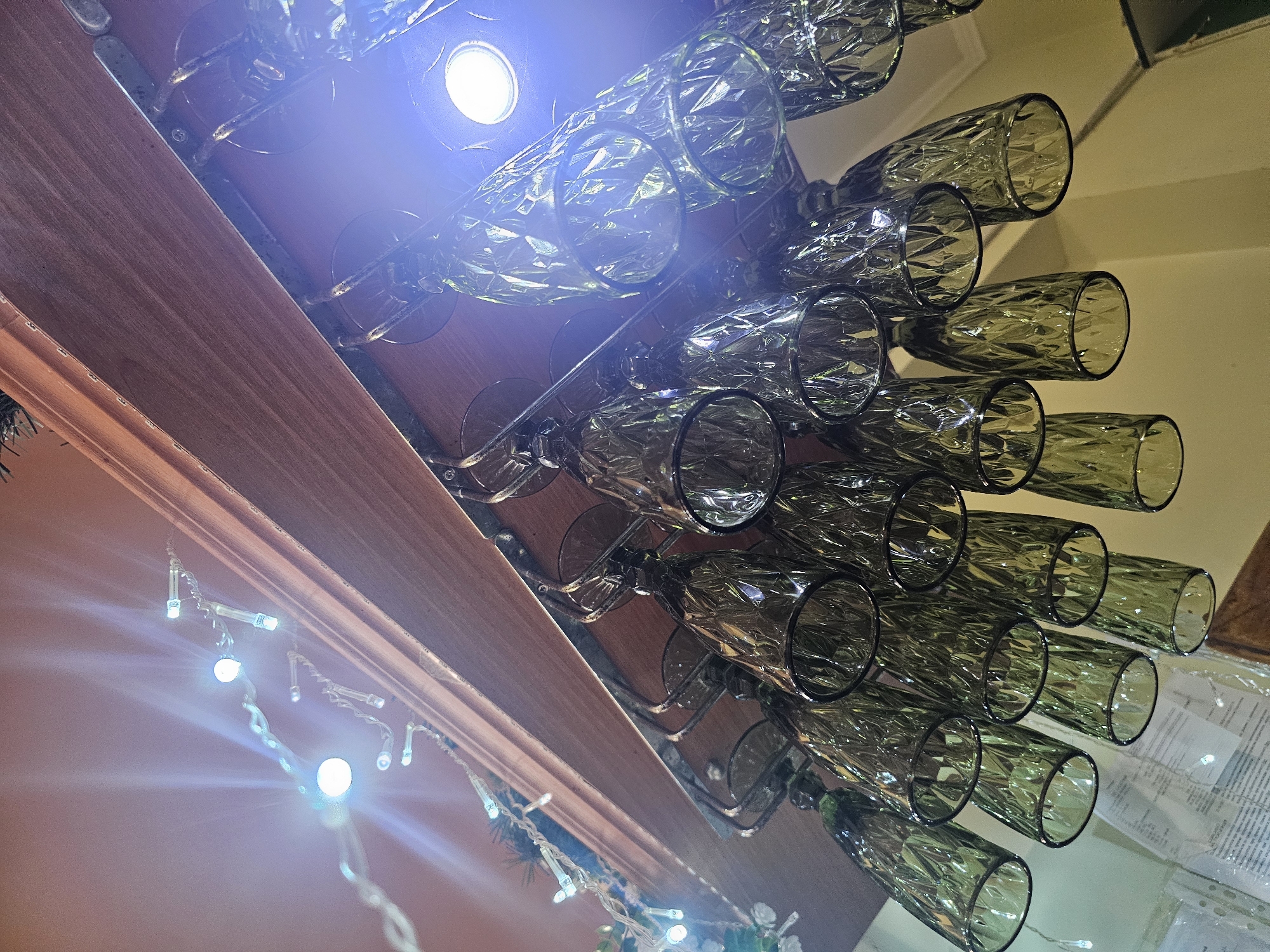 An upward-angle, eye-level shot captures a rack of green, faceted wine glasses hanging upside down from a wooden shelf. String lights with white bulbs are draped above the glasses, creating a festive atmosphere. A bright light source, possibly a bulb, is visible in the upper right corner, casting a strong blue-white glare. The background is blurred, but hints of a wall and possibly other shelving or decorations can be discerned. There are no people visible in the image. The setting appears to be indoors, possibly a bar or dining area. The specific location in Skadovsk, Ukraine, cannot be determined from the visual information provided.