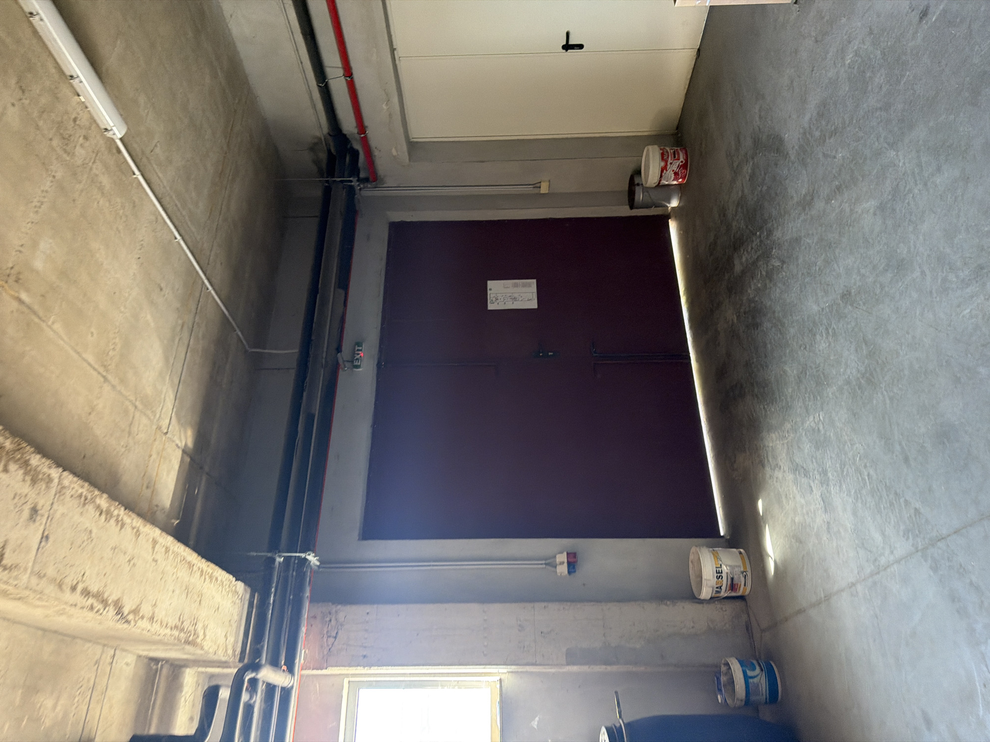 The image depicts an indoor concrete space, likely a storage area or garage, in Dolno Ablanovo, Bulgaria. The room features a large, dark red double door on the left and a white double door on the right. Above the dark red door, an "EXIT" sign is visible. The ceiling is unfinished concrete with exposed pipes and electrical conduits, including red pipes that run horizontally along the upper wall.
On the left side of the image, a window with light streaming through is present. Several industrial buckets are placed on the floor near the window and the red door. One bucket, labeled "MARSEL," is white with red and yellow markings. Another smaller blue and white bucket is next to it. A dark blue barrel is also partially visible on the far left.
The floor is made of rough, gray concrete. The lighting is dim, with some areas illuminated by sunlight from the window. No individuals are present in the scene, and no activities are being performed. The environment appears functional and utilitarian, with a focus on storage and access.