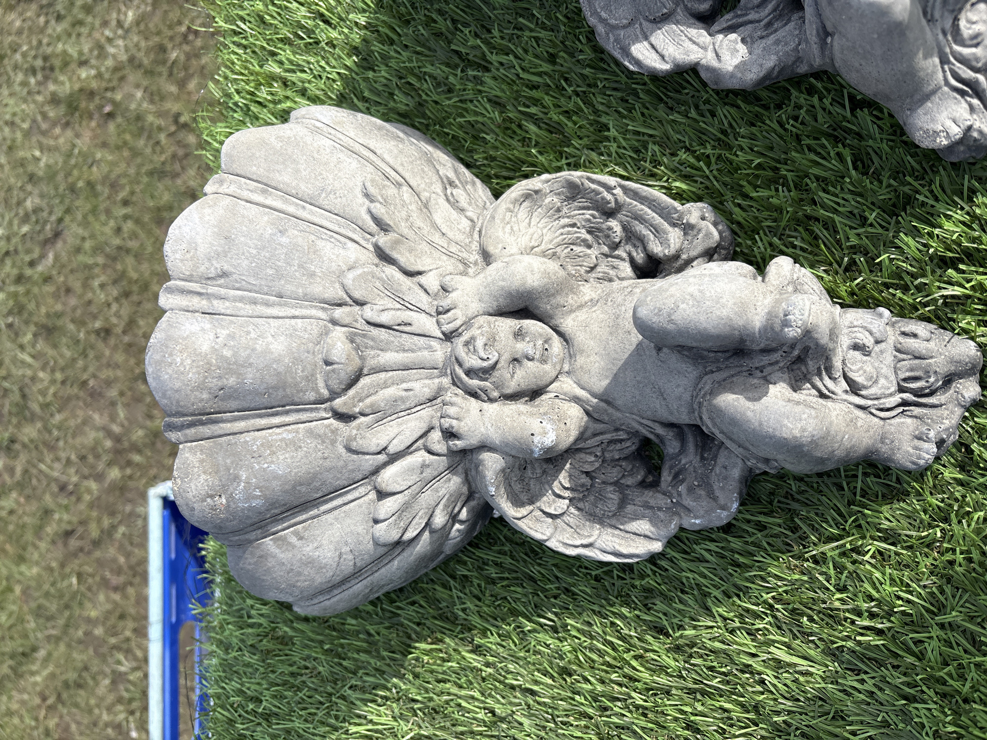 The image shows a stone angel statue lying on artificial green grass. The statue has a stylized shell or fan behind its back and another angel statue is partially visible in the upper right. A blue plastic container is visible to the left. The location is Chirk, United Kingdom.