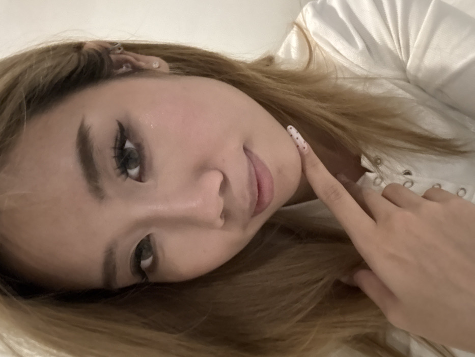 The image is a close-up, headshot-style portrait of a young adult female. She has light brown/blonde hair, styled straight and parted, framing her face. Her eyes, possibly enhanced by grey or hazel contact lenses, feature prominent dark winged eyeliner and long, dark eyelashes. Her eyebrows are well-defined. She has fair skin, with a subtle blush visible on her cheeks, and her lips are closed, forming a slight, subtle smile.
The subject is posed looking directly at the camera, with her right hand (viewer's left) raised. Her index finger rests lightly against her chin or lower lip, with the thumb and other fingers partially visible. Her fingernails are elongated, shaped, and painted white with small, dark polka dots. She is wearing a white garment, possibly a top or shirt, visible from the neck down, featuring a central lacing or eyelet detail. Accessories include a small, circular, silver-colored stud earring in her right earlobe (viewer's left), and a small hoop earring in the cartilage of the same ear.
The background is indistinct and appears as a plain, light-colored surface, suggesting an indoor setting. There are no discernible environmental details or objects in the background to indicate a specific city or location, therefore, the city of Yuzhong District, China, cannot be determined from this image.