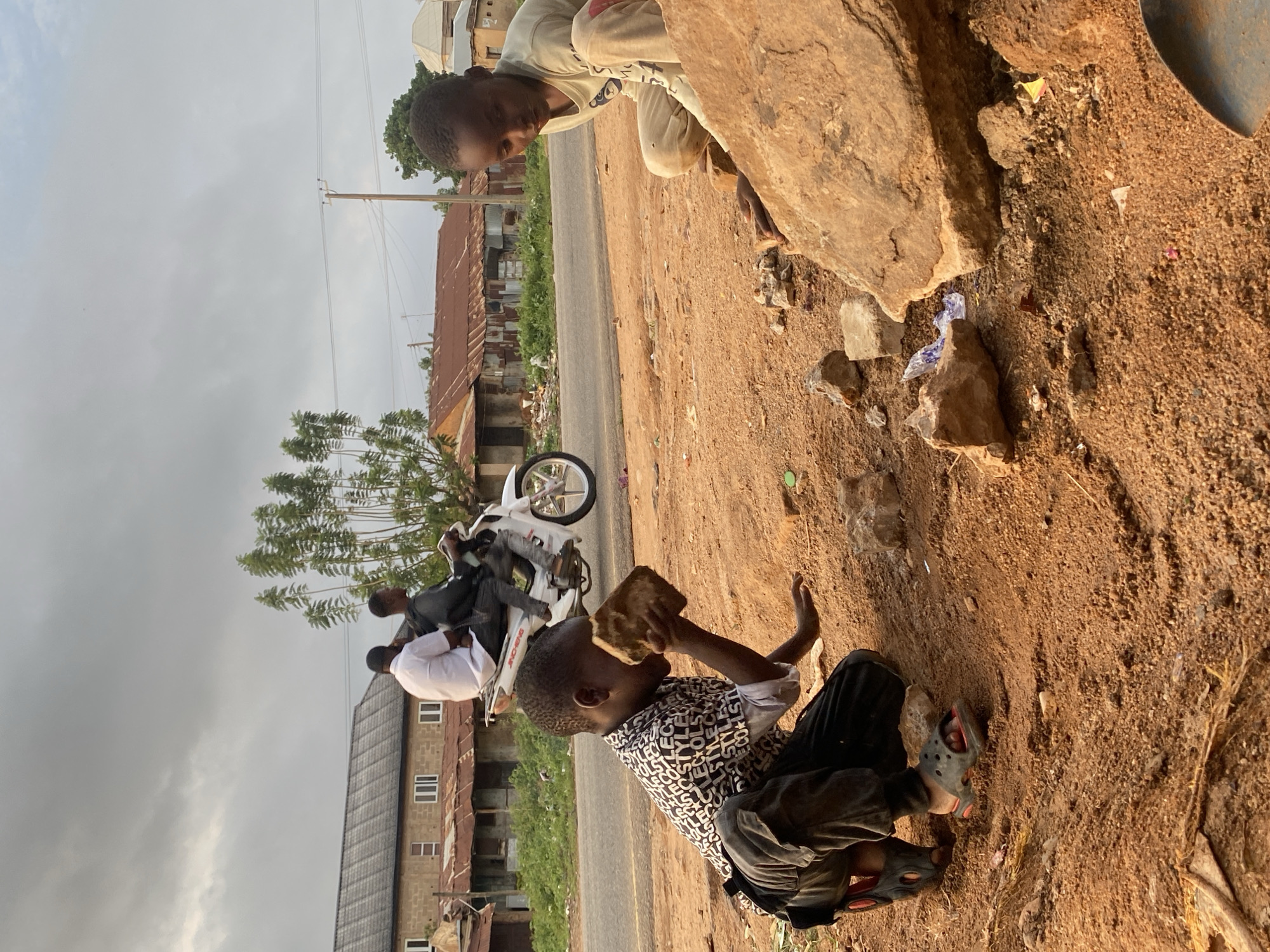 Two boys play by a roadside in Vom, Nigeria. One boy kneels, holding a chunk of earth to his face. The other boy, in a light-colored shirt, peers down from behind a large rock. A motorbike with two passengers is visible on the adjacent road. Buildings line the street, and the sky is overcast. Litter is scattered on the ground near the rocks.