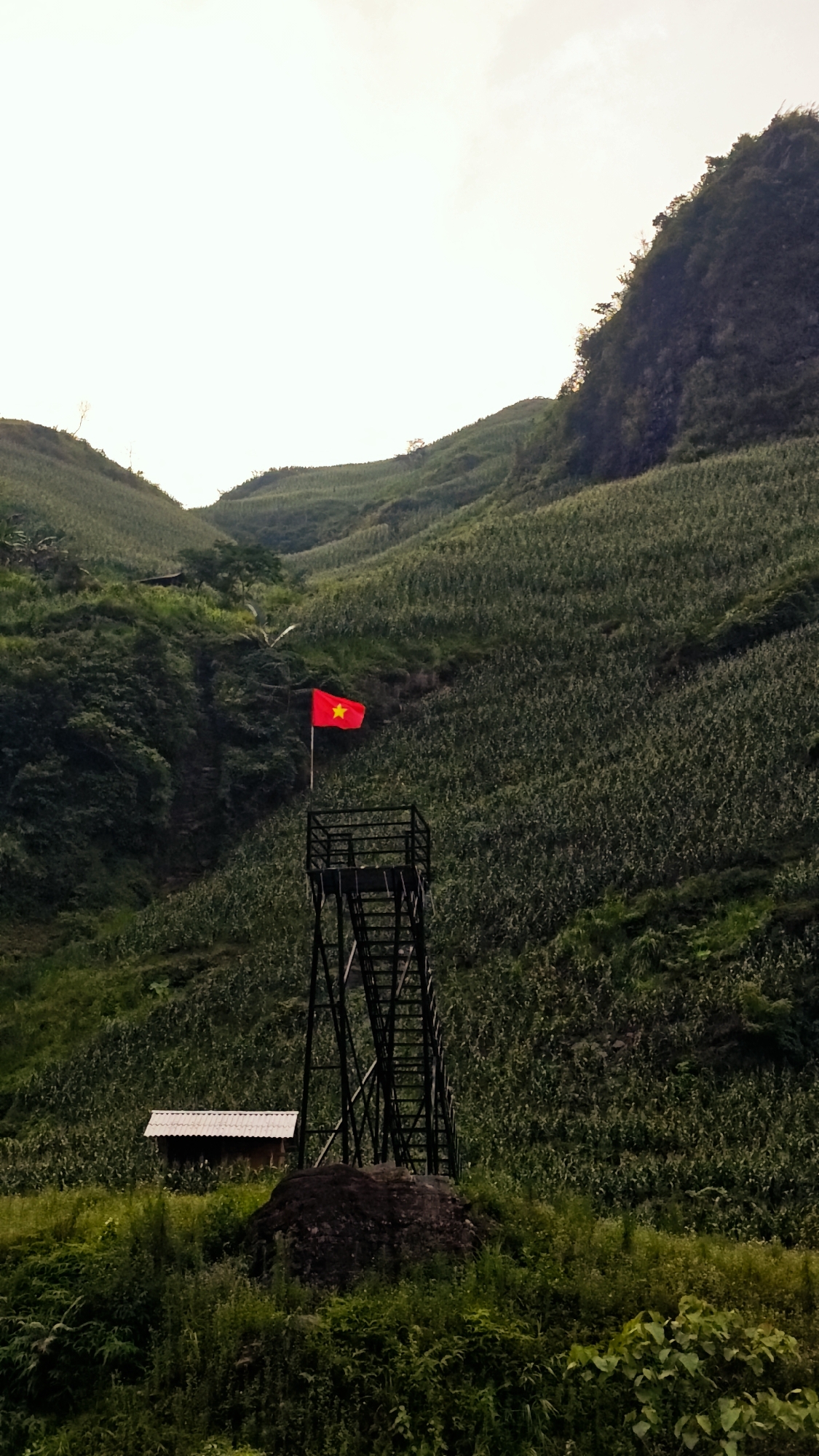 The image depicts a rural landscape in Du Già Commune, Vietnam. The scene is dominated by rolling green hills covered in dense vegetation, likely crops. A metal observation tower stands prominently in the foreground, topped with a red Vietnamese flag bearing a yellow star. A small, simple building with a corrugated roof is situated near the base of the tower. In the foreground, there are dense green plants and a dark-colored rock. The sky is overcast and bright, creating a soft light. No people are visible in the frame.