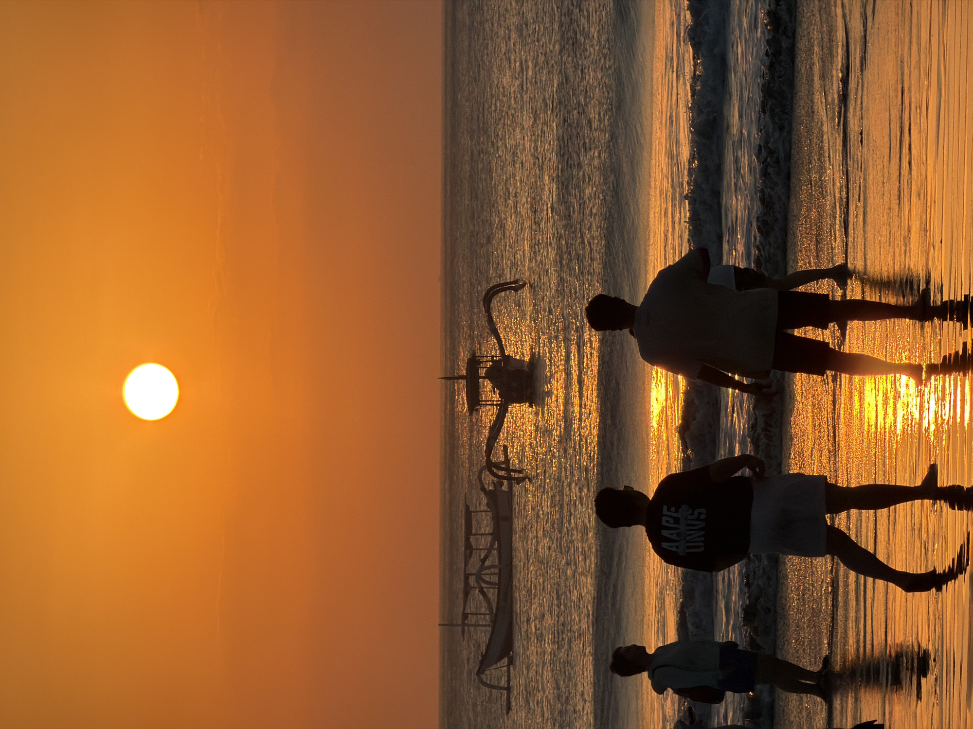 This is a vertical, low-angle photograph taken at sunset in Kuta Selatan, Indonesia. The scene depicts a beach with two traditional outrigger canoes on the water. Several silhouettes of people are visible standing on the wet sand near the shoreline.
The sun, a bright white orb, is low in the sky on the left side of the frame, casting a warm, orange glow across the scene and reflecting off the water and wet sand. The ocean appears calm with gentle waves. The overall atmosphere is serene and peaceful, typical of a beach sunset.
One of the figures in the foreground is wearing a dark t-shirt with white text that appears to read "AAPE UNVS". Another figure is wearing a light-colored hoodie. The figures are standing, and their reflections are visible on the wet sand. The presence of the canoes and the tropical sunset strongly suggest a location in Southeast Asia, consistent with Kuta Selatan, Indonesia.