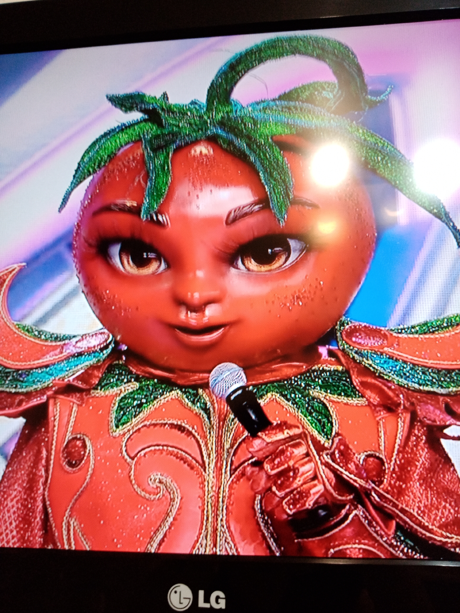 The image shows a person in a "Tomato" costume, part of the "The Masked Singer" show. The costume includes a red, tomato-shaped head with green leaf-like extensions on top. The person is holding a microphone. The costume has red and green patterned clothing with gold accents. The image is displayed on an LG television screen. There is no evidence this image was taken in Elektrostal, Russia.