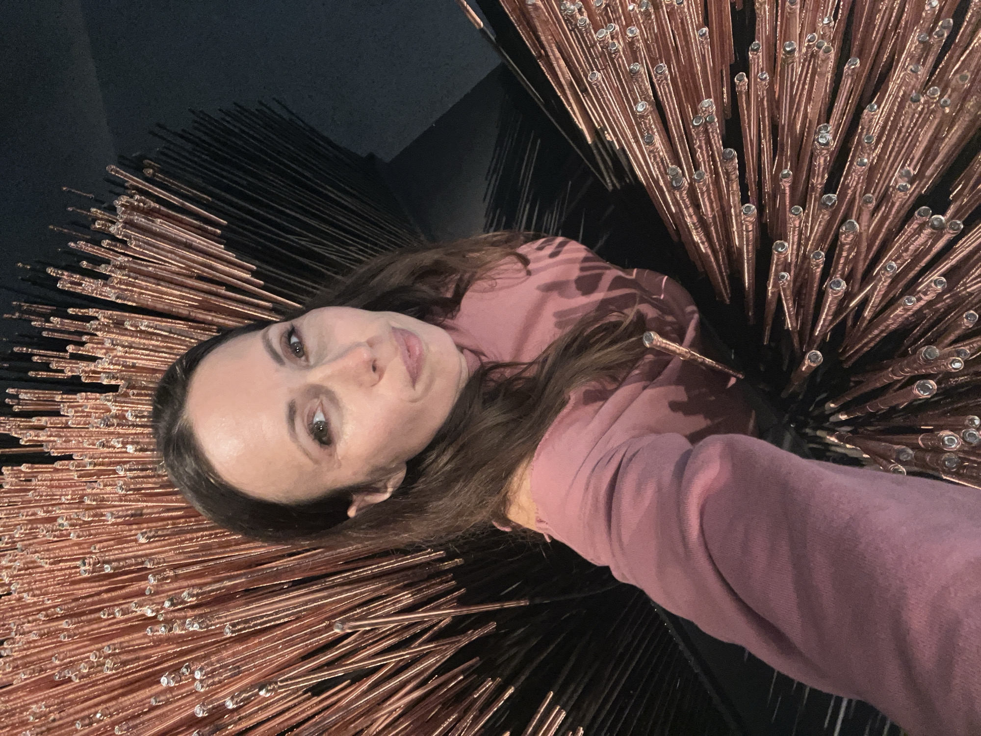 A woman is pictured in a selfie surrounded by an art installation comprised of copper rods capped with clear spheres, possibly glass, that resemble raindrops. The installation appears to be circular, radiating outwards from a central point. She is wearing a pink hooded sweatshirt and has brown hair. Dark interior elements, possibly walls and the base of the installation, are visible in the background. The precise location in Moscow, Russia, cannot be determined from the image.