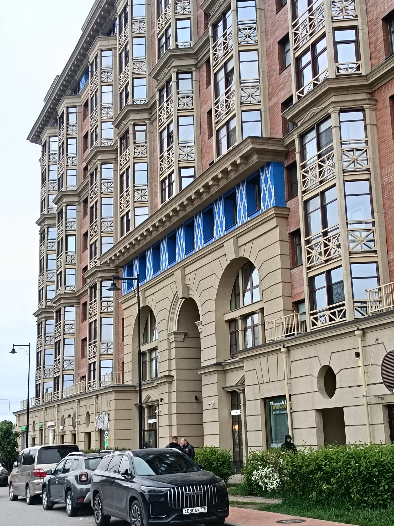 The image depicts a multistory building in Saint Petersburg, Russia. The architectural style features brick construction with prominent window bays, decorative woodwork, and a facade with arches, blue panels, and classical detailing. Several parked vehicles line the street in front of the building, including a dark-colored SUV with license plate "A080AX778." Two people stand near the entrance, and another figure is partially obscured by bushes closer to the building. Streetlights are visible on the left side of the image, and signage is present on the building's facade, including one that says "ПИВОМ." The sky appears overcast.