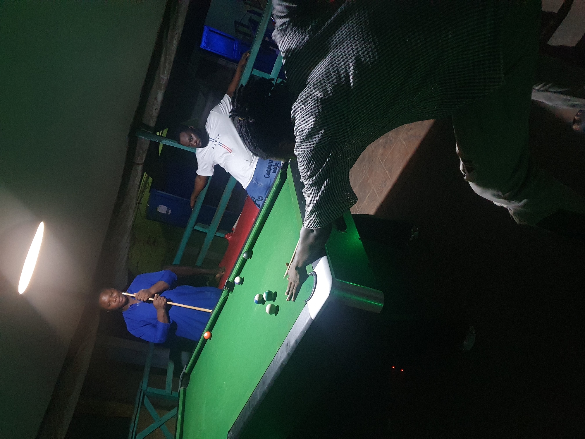 In Lokoja, Nigeria, three individuals are engaged in a game of pool on a green felt table under a bright overhead light. One person, wearing a blue top, stands behind the table, holding a cue stick. Another person, dressed in a white t-shirt and blue shorts, leans against a railing to the right of the table, observing the game. A third individual, in the foreground and facing away from the camera, is in the process of taking a shot, with their hand on the cue stick. Several pool balls are scattered across the table, including a red ball near the top left corner and a cluster of balls in the center. The environment appears to be an indoor recreational area with wooden railings and a dark background.