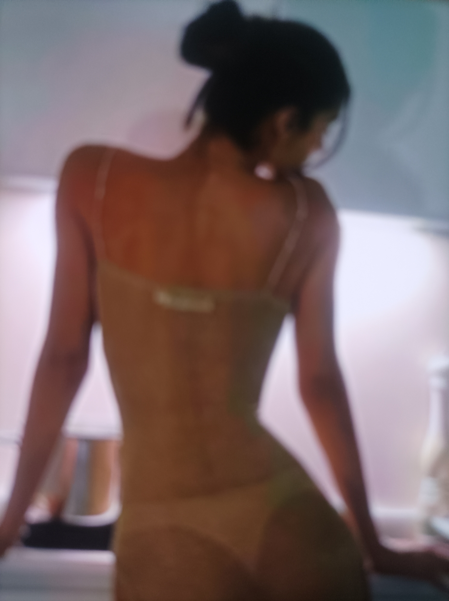 A blurry, medium shot captures the back of a person with dark hair pulled up in a bun. They are wearing a light brown, form-fitting garment that appears to be a leotard or bodysuit. The garment has thin spaghetti straps. The person is leaning forward slightly, with their left arm resting on a surface. The background is indistinct and appears to be an indoor setting with soft lighting. There are some blurred objects in the background, including what might be kitchen appliances on the left. The overall lighting is soft and diffused, creating a warm and intimate atmosphere. The image is of poor quality with significant blur and grain.