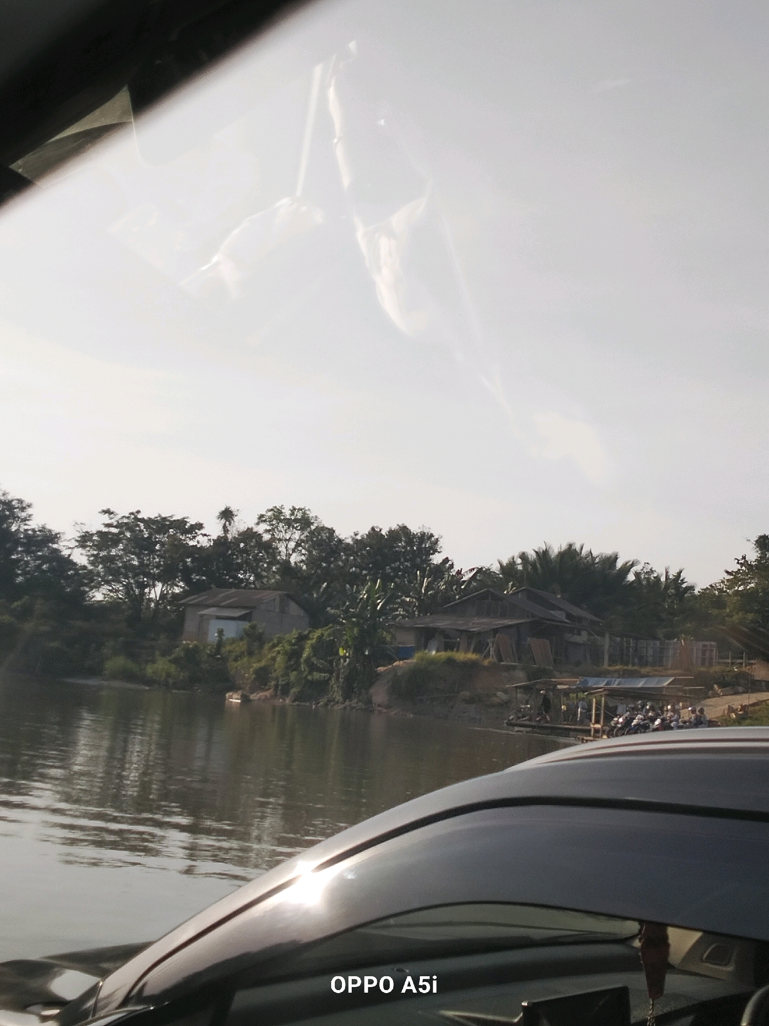 The image captures a view of a river and its surroundings in Tabanggele, Indonesia. In the foreground, the hood of a dark-colored car is visible, reflecting sunlight. The river occupies the middle ground, with calm water showing reflections of the sky and the opposite bank. On the far bank, a collection of buildings, some appearing to be houses and others more like sheds or workshops, are nestled amongst lush green trees. There are palm trees and other dense vegetation. A small wooden pier extends from the bank, with several people gathered and a line of motorcycles parked. The sky is bright and appears to be partly cloudy. The overall scene suggests a rural or semi-rural setting along a waterway.