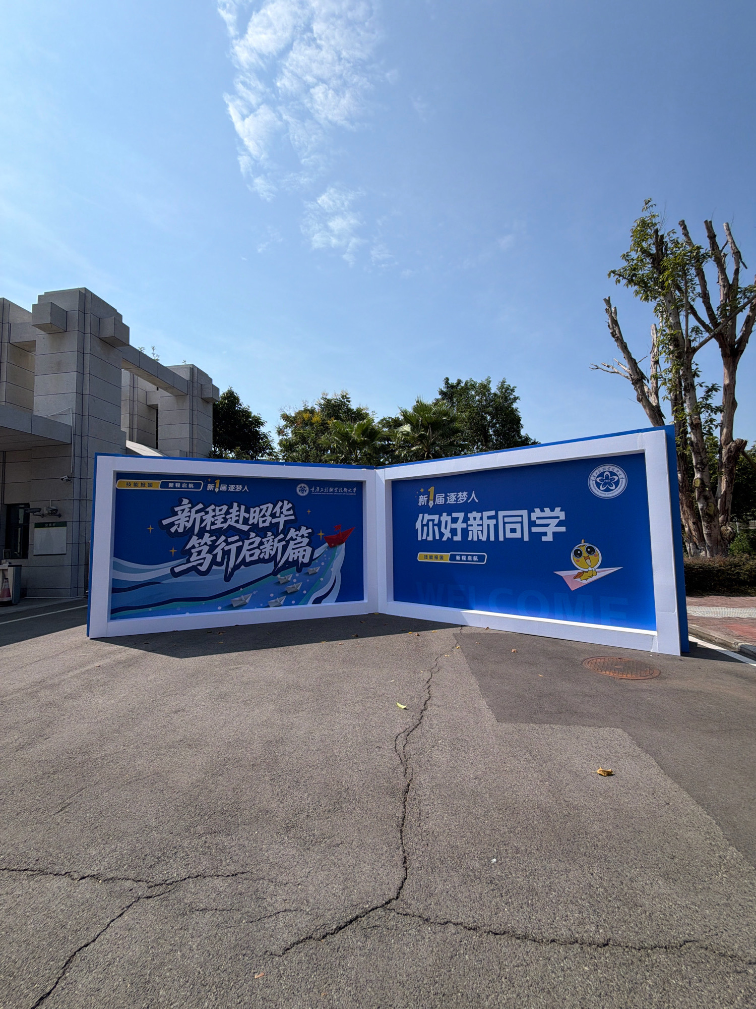 The image shows two large blue and white banners placed outdoors on an asphalt surface. The banner on the left has Chinese characters that translate to "New Journey, Blooming Dreams" and "Embark on a New Chapter". It features a graphic of a red paper boat sailing on a wave towards other smaller paper boats. The banner on the right has Chinese characters that translate to "Hello, New Classmate" and includes a cartoon chick riding a paper airplane. A circular emblem with Chinese characters is also visible on this banner. Behind the banners, a modern building with stone facade and trees can be seen under a clear blue sky with scattered clouds. The banners appear to be welcoming new students to an educational institution. The scene is likely located in Jiangjin District, Chongqing, China, given the presence of Chinese language text and architectural style commonly found in Chinese institutions. The ground shows cracks in the asphalt and some scattered leaves.