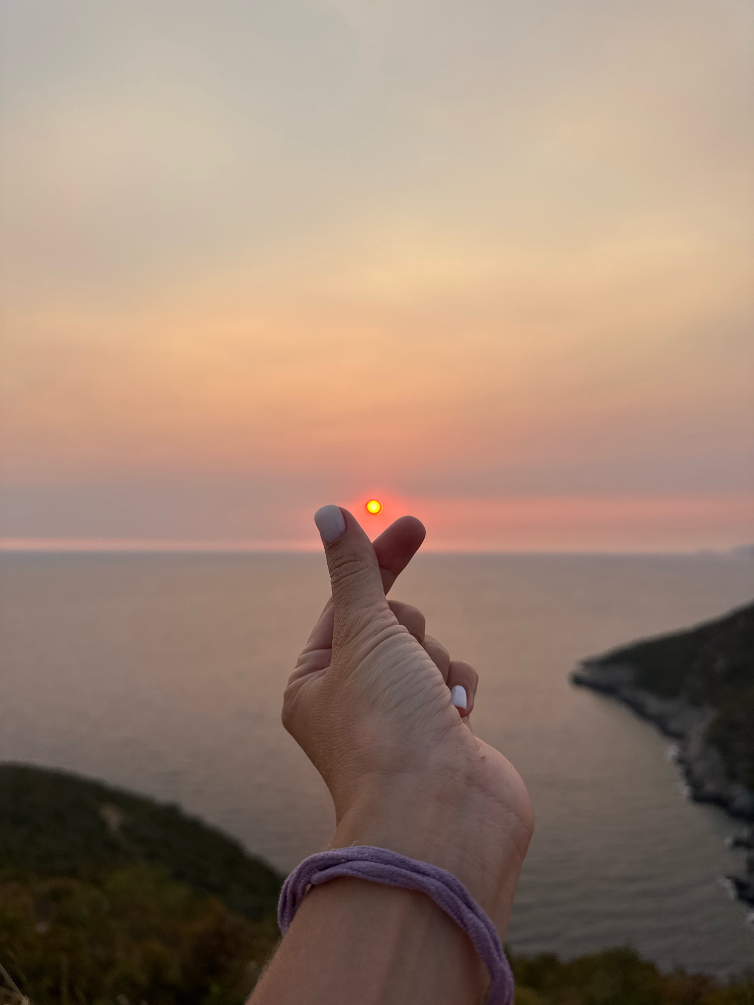 A close-up shot focuses on a person's hand, fingers forming a heart shape, with the sun, appearing as a small, bright yellow circle, positioned precisely between the thumb and index finger. The hand is adorned with a light purple bracelet around the wrist. The background reveals a serene seascape at sunset. The horizon line is clearly defined, separating the calm, muted orange and pink hues of the sky from the darker, tranquil waters. To the right, a rugged, tree-covered coastline juts out into the sea, its shape softened by the atmospheric light. The foreground consists of the upper part of the person's arm and wrist, and hints of green foliage on a hillside. The overall scene evokes a sense of peace and romance, with the hand gesture playfully framing the setting sun. This image was captured in the vicinity of the city of Zagora, Montenegro.