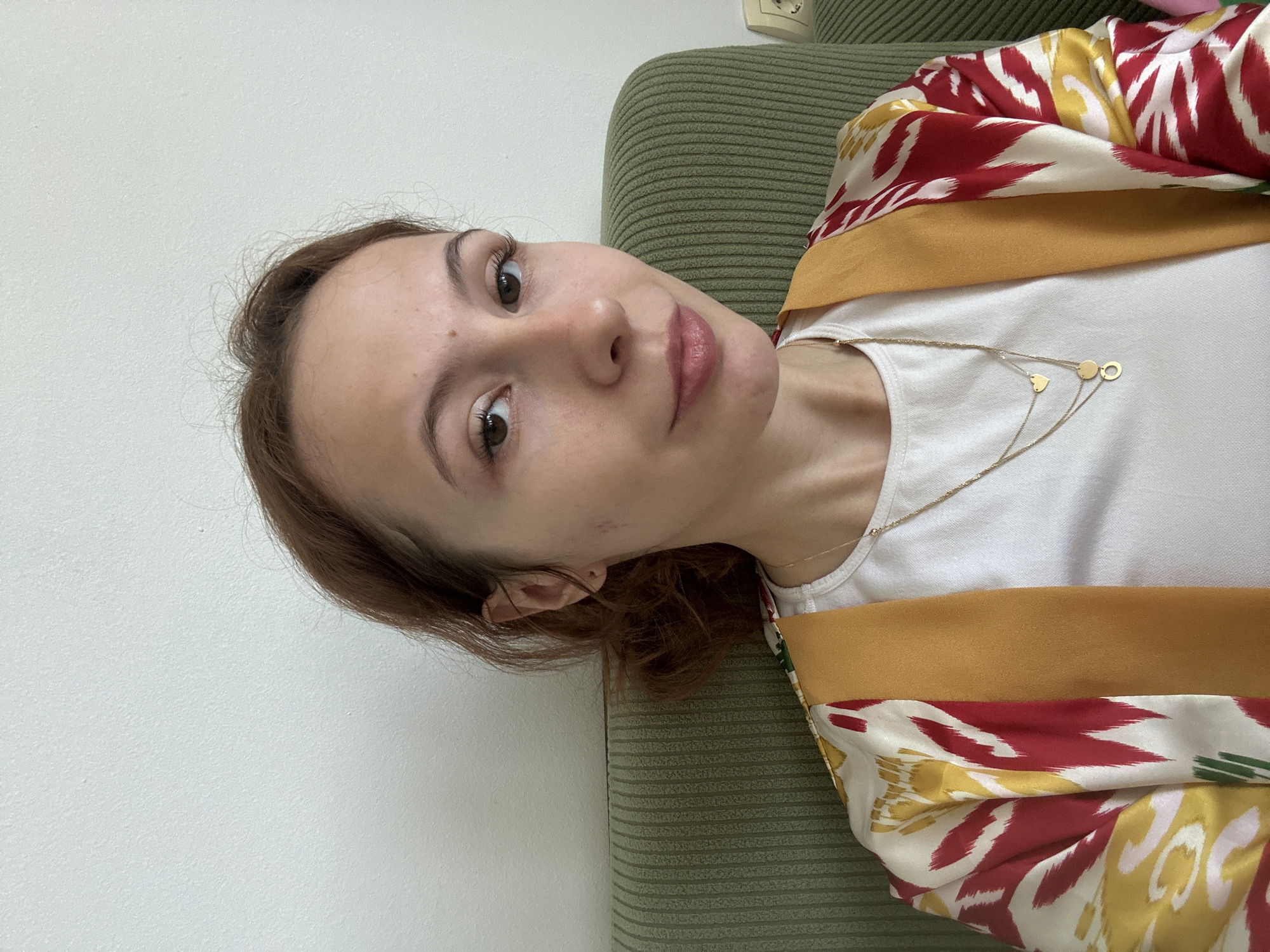 A woman with light brown hair is reclining on a green couch. She is wearing a white t-shirt and a patterned kimono with red, yellow, and white designs. A delicate gold necklace adorns her neck. The background consists of a plain white wall. The photo was taken in Novi Sad, Serbia.