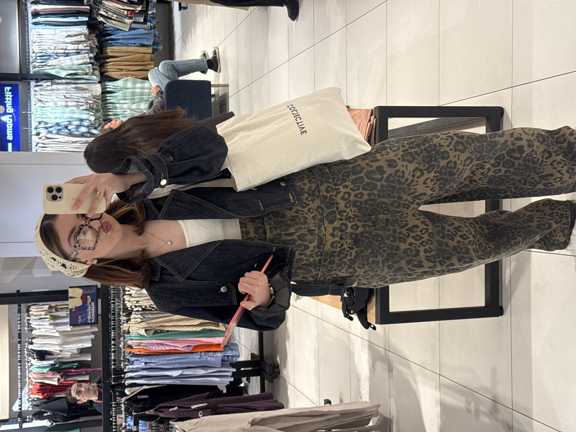 A woman is taking a selfie in what appears to be a clothing store. She is wearing a white top, denim jacket, and leopard print pants. She also has glasses and a crochet headband on. She is holding a phone in one hand and a pink item in the other. Behind her are racks of clothes. The store has a white tiled floor. The text on the bag reads "COEJCLIAE". The location and city are not discernible from the provided visual information.