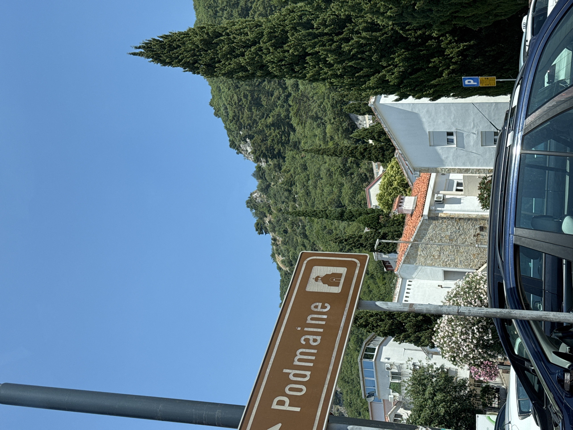A street sign with white text on a brown background reads "Podmaine" and depicts a small church symbol to the right. An arrow on the left side of the sign points to the left. The sign is attached to a dark gray pole. In the background, there are white buildings with red tile roofs, lush green trees, and a rocky, tree-covered hill under a clear blue sky. A tall, slender evergreen tree is positioned to the right of the sign, and other trees and foliage are visible throughout the scene. Several cars are parked in the foreground, including a dark blue SUV and a white car. A yellow parking sign with a blue 'P' is visible on the right side of the image, and a white taxi is parked behind the blue SUV. The overall scene appears to be a sunny day in a coastal town, likely Budva, Montenegro, given the characteristic architecture and vegetation.