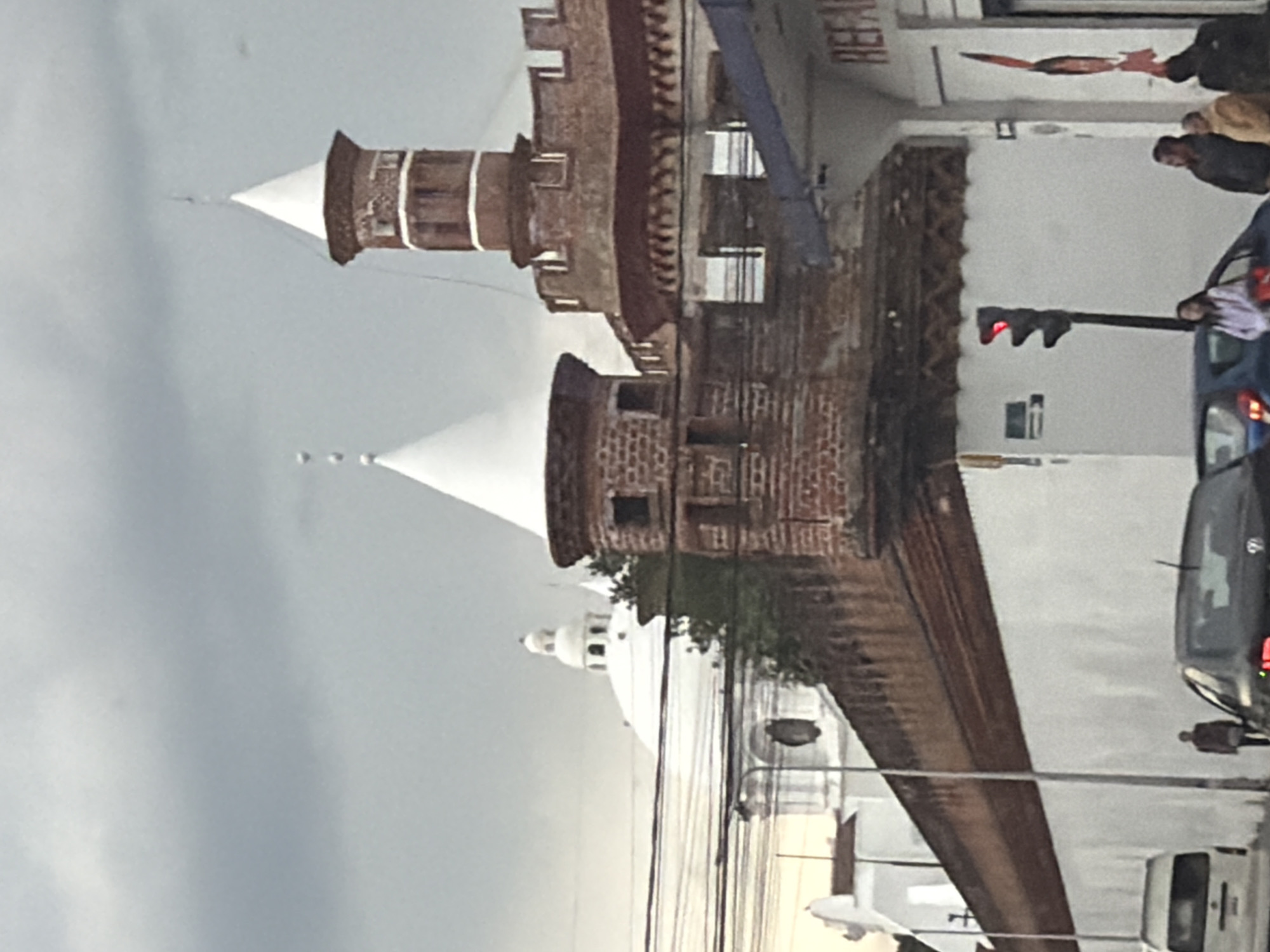The image depicts a street scene in Puebla, Mexico, featuring distinctive architectural elements. Dominating the view are two towers with white conical tops, connected to a building with red brick and white walls. Adjacent to the building is a city street where cars are stopped at a red traffic light. Several people are visible on the sidewalk near the street. Overhead electrical wires stretch across the sky, which is overcast with a gray sky. In the distance, a white domed structure is visible.