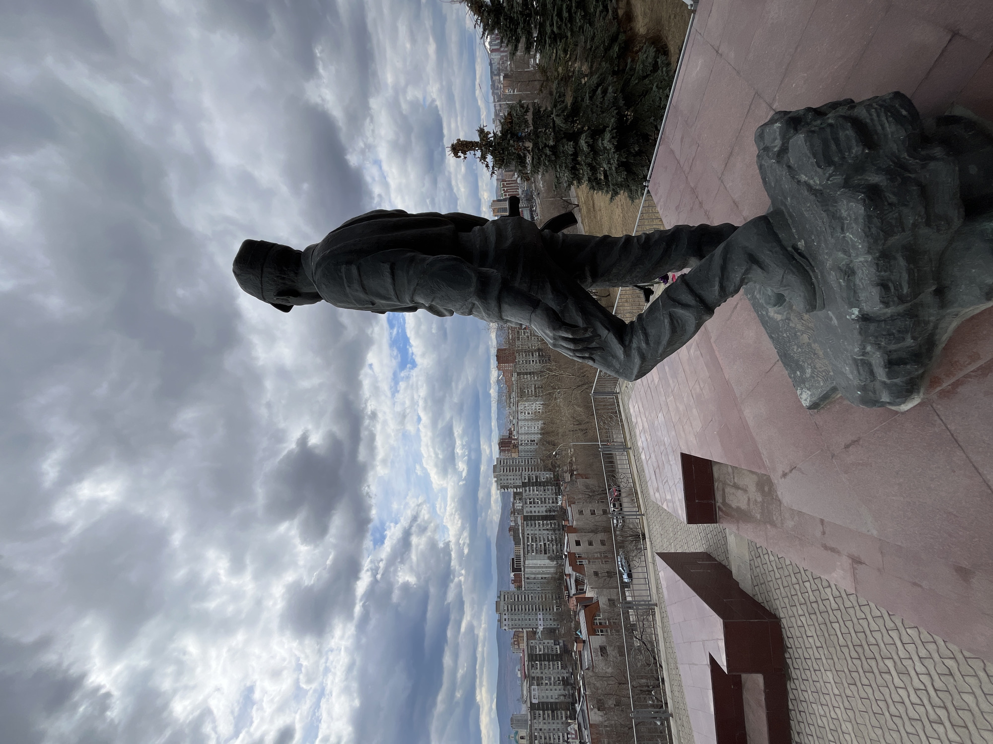 An outdoor scene captures a large, dark-colored statue of a man, likely a soldier or worker, standing on an elevated reddish-brown paved platform. The statue, depicted in practical attire and wearing a cap, faces away from the viewer, looking out over an extensive urban landscape. A rifle or similar implement appears to be slung across its back.
The setting is an overlook in Krasnoyarsk, Russia, providing a panoramic view of the city. Numerous multi-story apartment buildings dominate the mid-ground, stretching towards distant hills or mountains on the horizon. Clusters of evergreen trees are present near the statue, enhancing the memorial-like atmosphere. The sky is heavily overcast with prominent gray and white clouds, though occasional patches of blue sky are visible, indicating it is daytime.
The monument's base features angular, geometric structures made of the same reddish-brown material as the paving. A metal railing borders the platform, separating the monument from the city below. The overall impression is one of a solemn monument surveying the urban environment. No discernible text is visible in the image.