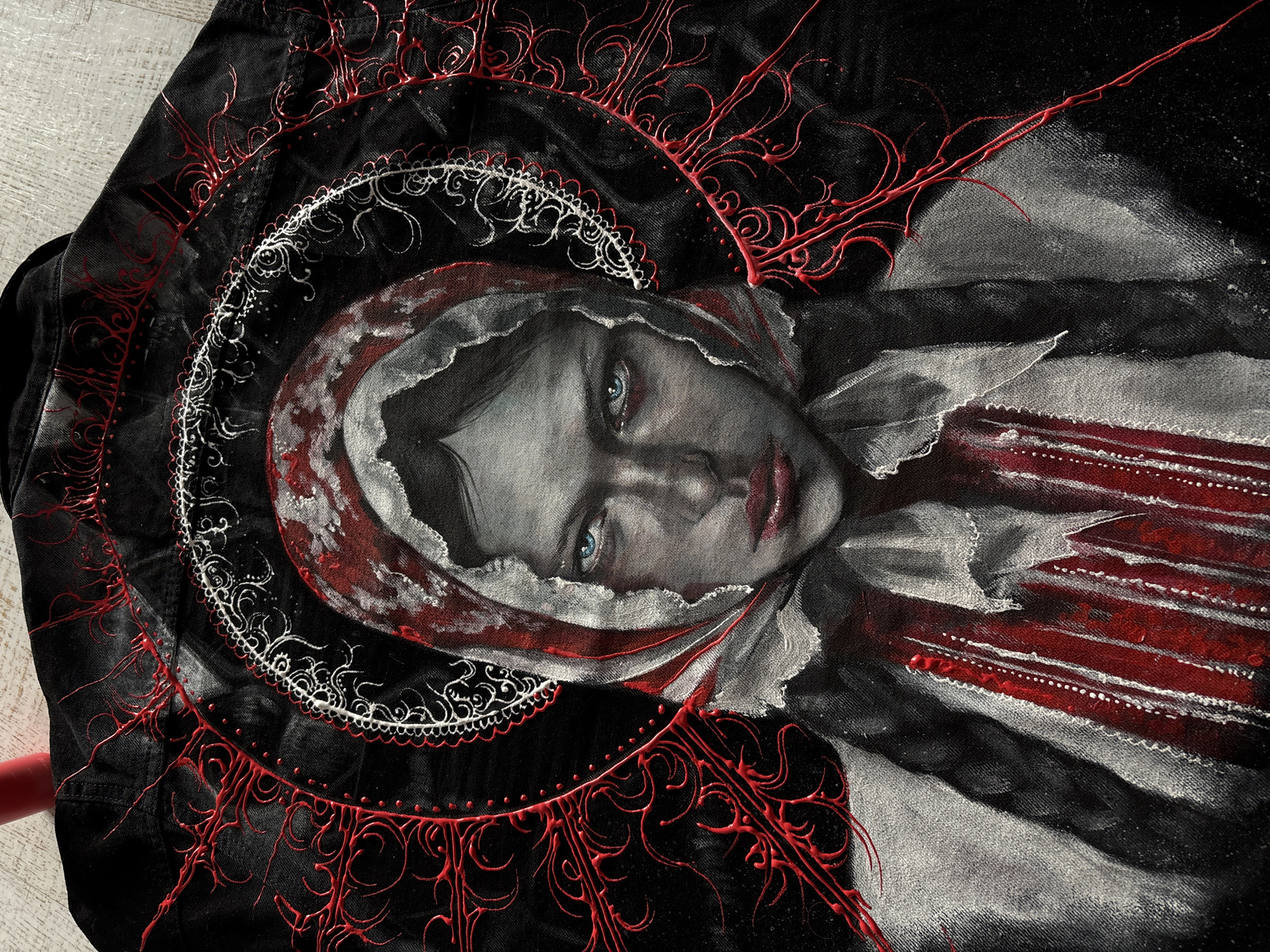 An intricately hand-painted portrait of a woman is displayed on a black fabric, likely a jacket, laid flat on a light-colored surface, possibly wood. The woman's face is rendered in grayscale tones with striking blue eyes and reddish lips, conveying an intense or melancholic expression. She is adorned with a reddish and white head covering, possibly a hood or shawl, and a ruffled collar.
The portrait is encircled by elaborate, halo-like patterns. An inner ring consists of delicate white, ornate scrollwork, while an outer, wider ring features raised, glossy red lines forming swirling, organic shapes that emanate outwards, creating a textured, almost three-dimensional effect against the black fabric. Additional red embellishments extend from the halo onto the jacket's sleeves and body. The overall color palette is dominated by black, white, red, and shades of gray, with the vivid blue of the eyes providing a focal point.
No specific activity is taking place beyond the presentation of the artwork itself. There is no visible text. The artwork appears to be a finished piece, possibly originating from Krasnoyarsk, Russia, given the provided context.