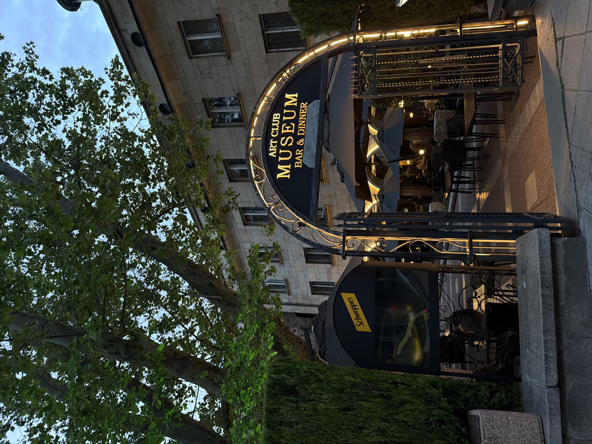 The image captures the exterior of "Art Club Museum Bar & Dinner" in Sofia, Bulgaria. The entrance is marked by an ornate, arched black sign with the establishment's name in gold lettering. The sign is illuminated with string lights. A decorative wrought-iron gate leads to an outdoor seating area with tables and chairs under umbrellas. A Schweppes sign is visible near the entrance. The restaurant is situated in front of a multi-story stone building with windows. Lush green trees and hedges surround the entrance, partially obscuring the building. The time of day appears to be evening or dusk.