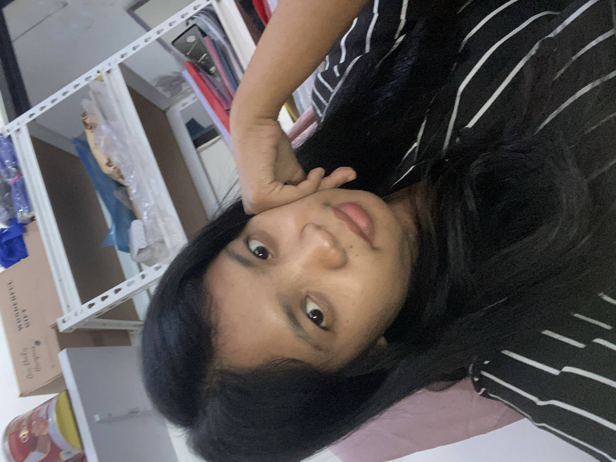 A person with dark hair and fair skin is pictured indoors. The individual is wearing a black and white striped shirt and resting their head on their hand. The background includes shelves containing various items, including clothes and boxes. The specific location in Negara, Indonesia is not discernible from the image.