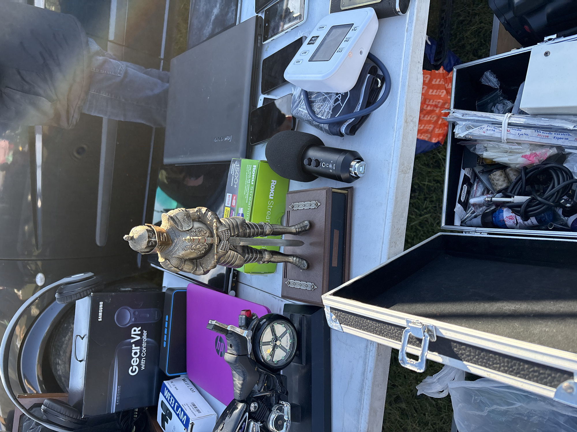 This image, likely captured in Chirk, United Kingdom, shows a table covered with various items, possibly at a flea market or a yard sale.
Objects on the table include a Lenovo laptop, several phones, a blood pressure monitor, a microphone, a Roku streaming device, a bronze figurine of a knight, a decorative book, a Samsung Gear VR headset, a webcam box, a purple HP device, a motorcycle toy with a clock, an opened metal case with items inside (needles, cables, etc.), and a pair of headphones.
The table is positioned outdoors, with evidence of grass and possibly a vehicle nearby. The person taking the photo appears to be standing next to the table, based on the partial view of clothing in the image.