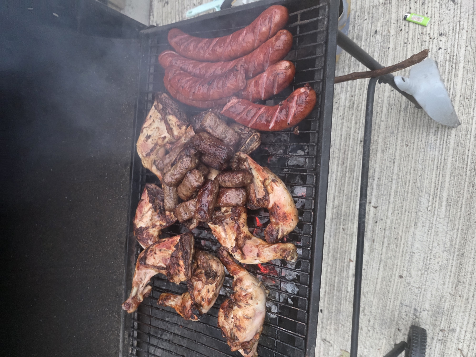 The image shows a barbecue grill with various meats cooking over coals. There are several pieces of chicken, likely leg quarters, some sausages, and what appears to be ribs or some other cuts of beef. The grill is positioned on a concrete surface. The environment is outdoors, with the dark interior of the grill lid visible at the top. There is smoke rising from the grill, indicating that the food is actively cooking. The scene appears to be a casual outdoor meal preparation. This image does not provide any specific details or context to determine its location within the city of Maynooth, Ireland, or any other specific city.