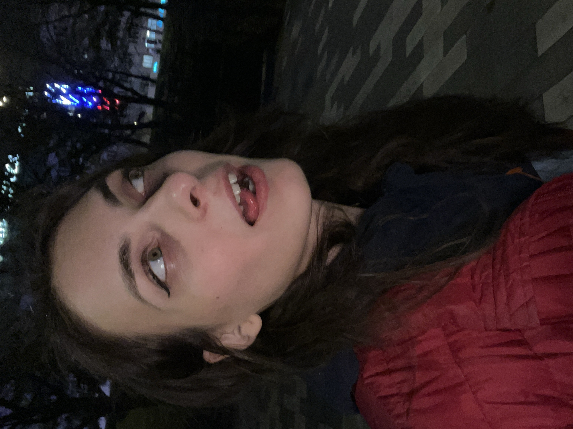The image is a selfie taken at night, depicting a woman with long, dark hair. She has fair skin, dark eyeliner, and her mouth is slightly open, revealing her tongue. She is wearing a dark blue garment and a red quilted jacket. The background shows blurry city lights and trees, suggesting an urban outdoor location. The style of the image and the presence of city lights indicate the location is likely within a city but the city of Krasnoyarsk, Russia is not clear from the photo. The angle of the selfie is slightly rotated.
