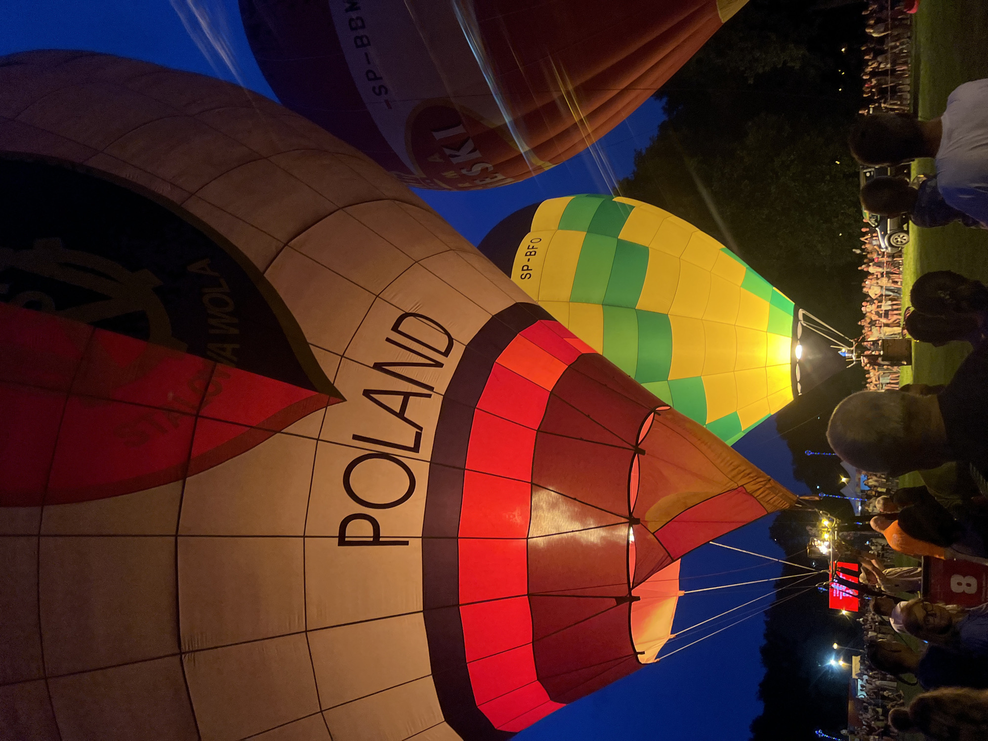 The image captures a night scene at a hot air balloon festival in Tychy, Poland. Several hot air balloons are illuminated, with the prominent "POLAND" text visible on one of them. The balloons are in various stages of inflation or preparation, with some tethered and others appearing ready for ascent. A crowd of people is gathered in the background, watching the spectacle. In the foreground, several individuals are looking towards the balloons. The atmosphere appears festive, with artificial lights illuminating the area. The environment is an open field with trees forming a dark backdrop against the night sky.