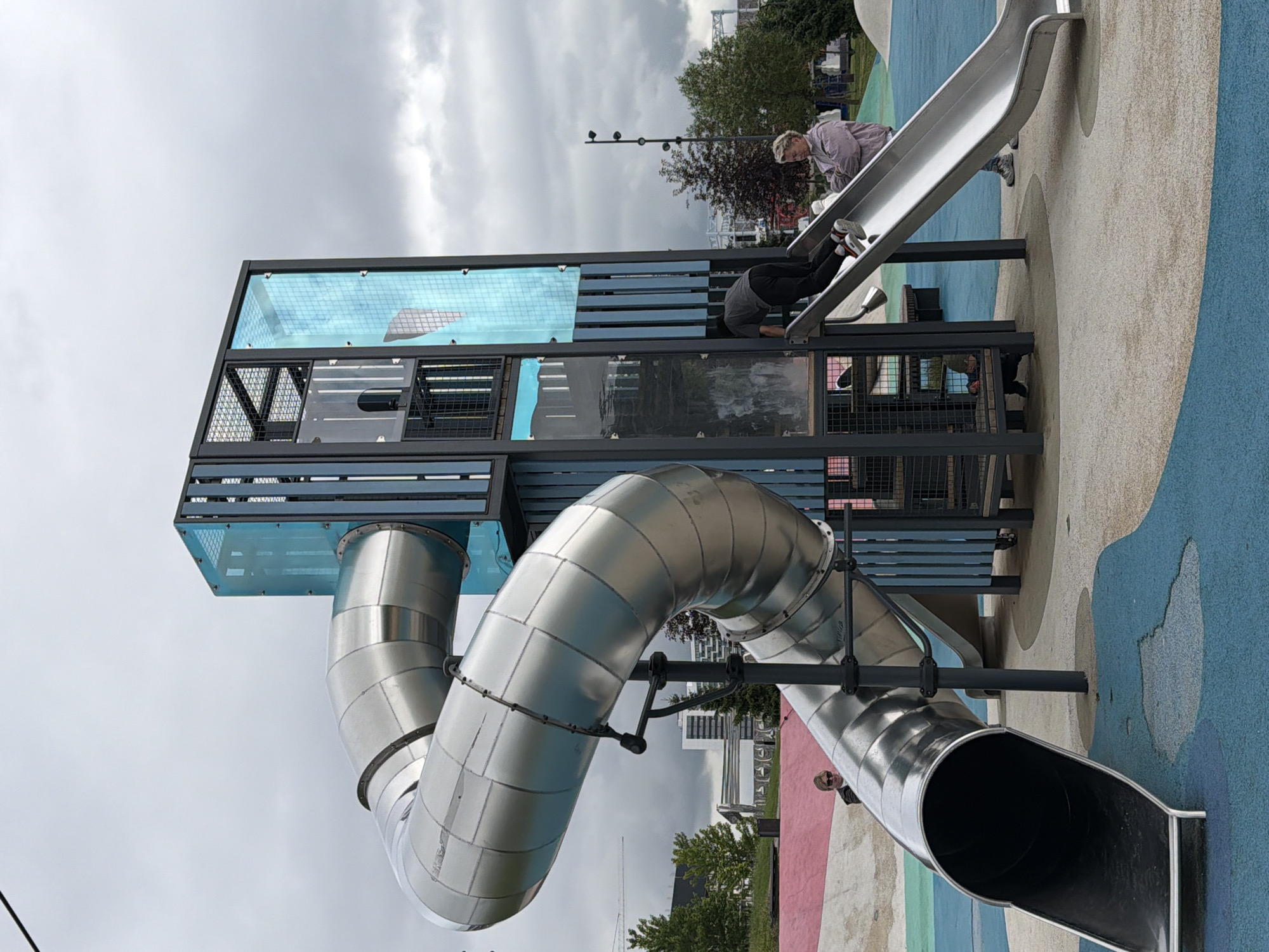 The image captures a playground scene on an overcast day in Khimki, Russia. The primary focus is a modern, multi-level playground structure featuring a large, silver, tubular slide that curves downwards. Two individuals are visible on the playground. One person, wearing a light-colored jacket and dark pants, is in a crawling or descending position on a silver slide at a higher level of the structure. Another person, wearing a grey shirt and dark pants, is also on a slide, appearing to be on the same structure.
The playground has a brightly colored, patterned surface beneath it, with sections of blue, beige, and pink. The playground structure itself is a blend of metal, glass panels with a blue tint, and dark framing, suggesting a contemporary design. In the background, trees and buildings are visible, hinting at an urban or suburban setting. The sky is cloudy, indicating diffused light and possibly cooler weather. The overall mood of the image is one of playful activity, despite the overcast conditions. There is no visible text in the image.