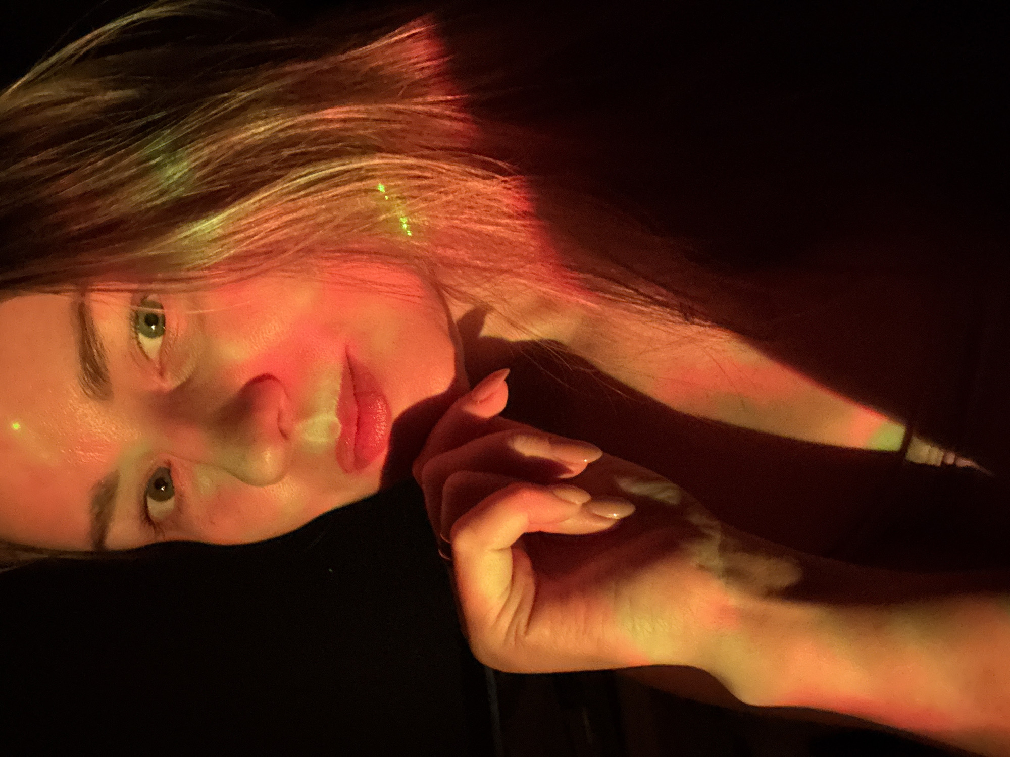 A young woman with light skin and long, blonde hair lies on her side in a dark environment illuminated with warm, reddish-orange light and a small amount of green light. Her head is slightly tilted upwards, and her hand is resting near her face. The woman has light-colored eyes, full lips, and manicured nails. Due to the absence of recognizable landmarks or indicators, it is impossible to determine whether or not this image was taken in Chaikovsky, Russia.